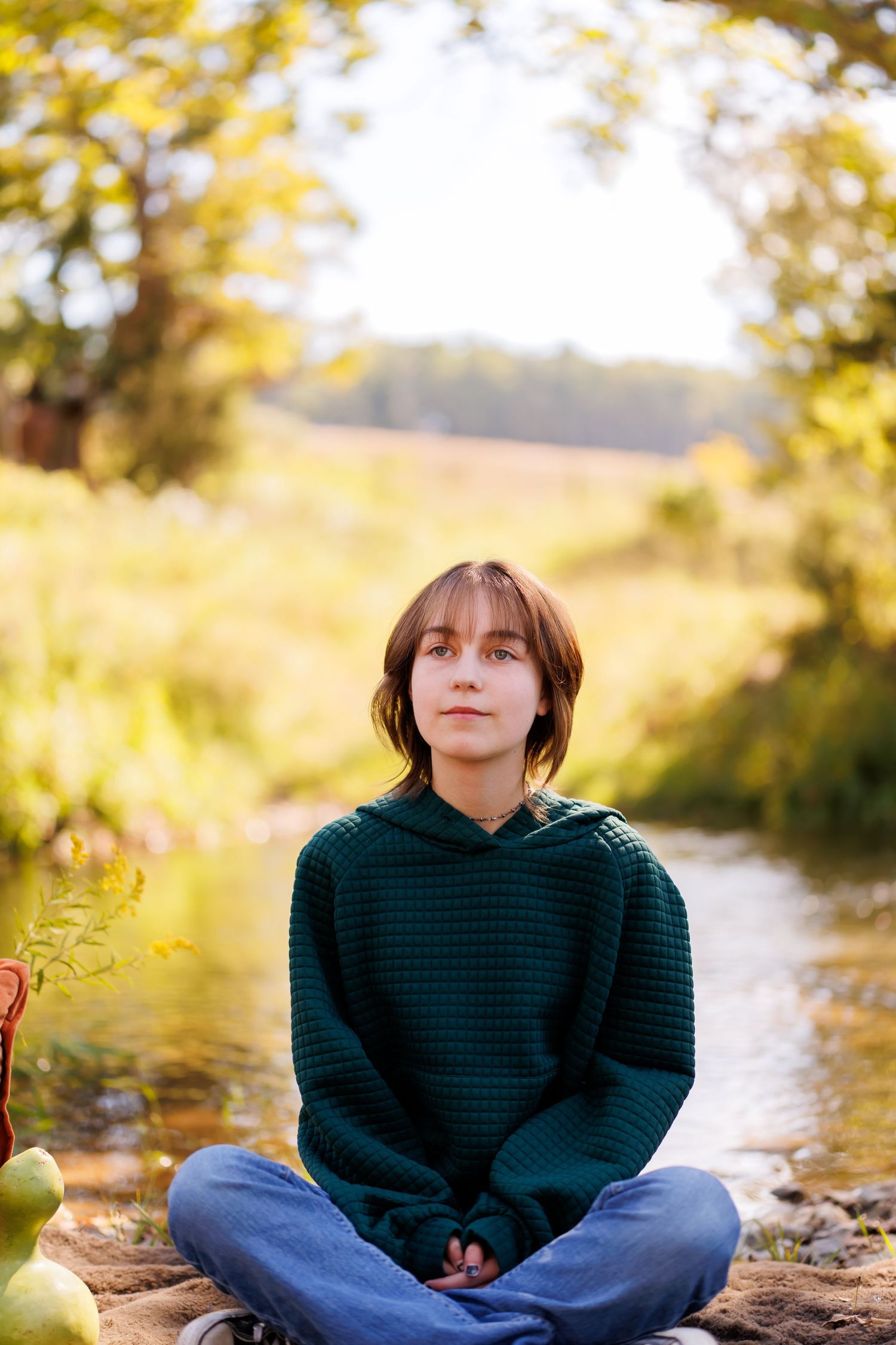 Teen girl in green sweater sitting cross-legged outdoors by a stream in a sunny autumn forest setting.
