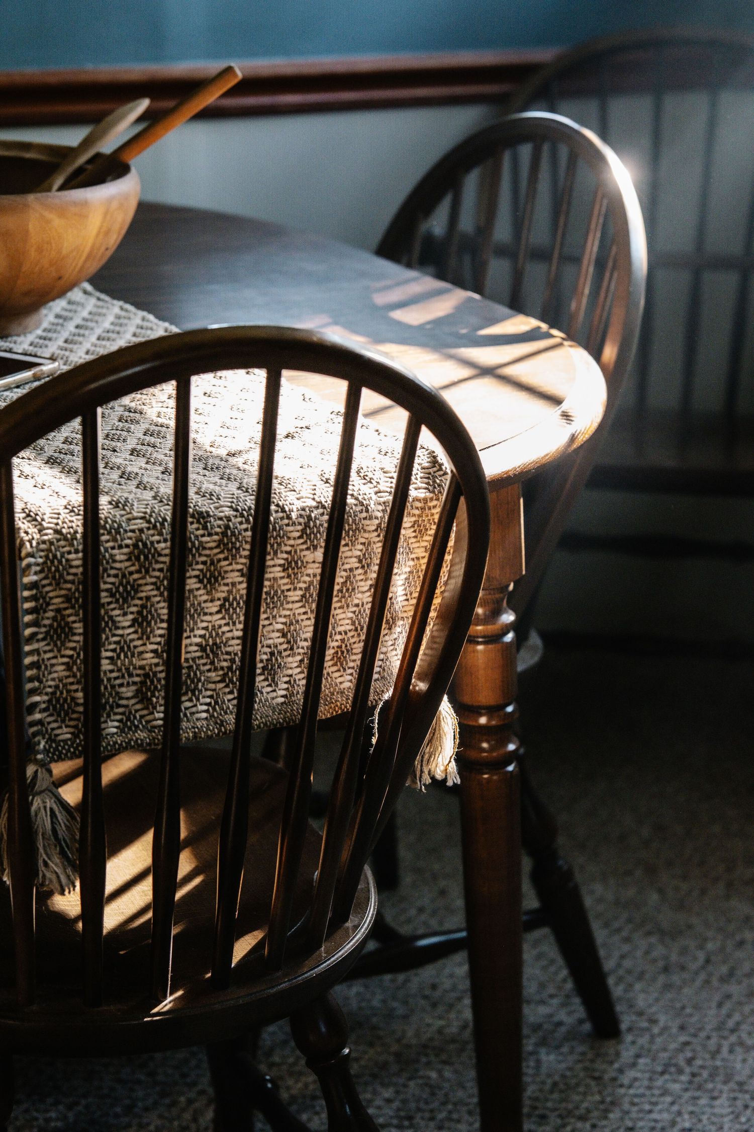 Vintage Windsor dining chair beside a dark wood table with woven table runner and wooden bowl in warm sunlight.