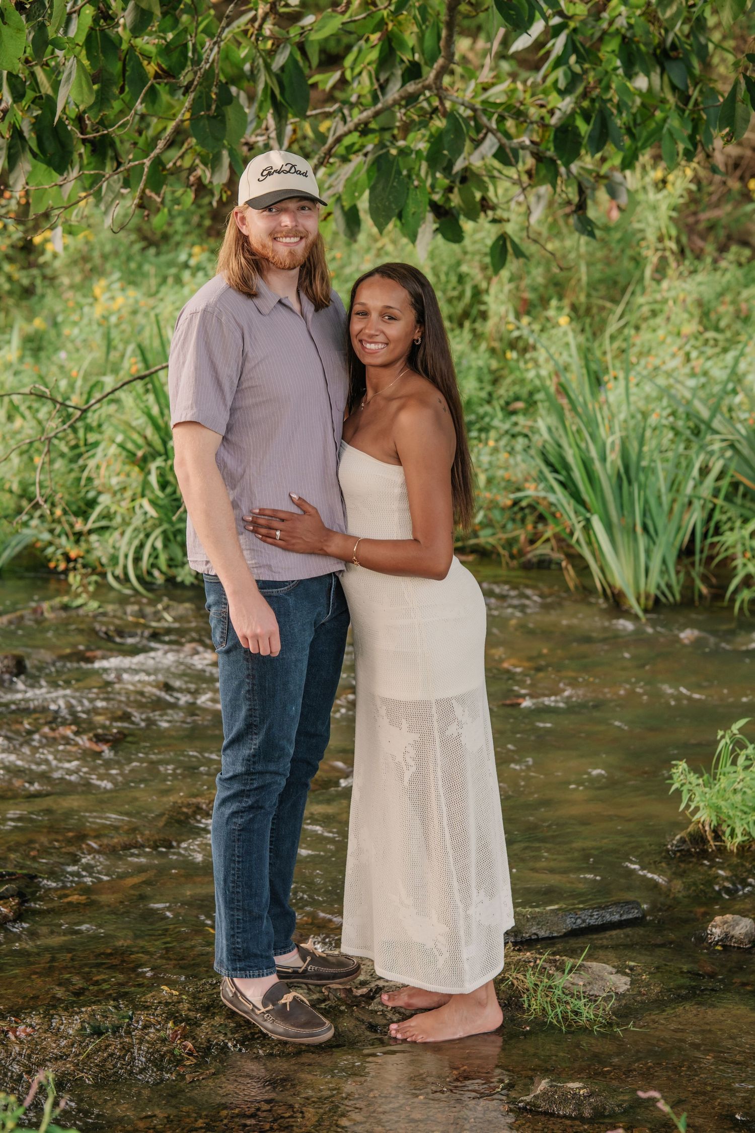 Couple standing in a shallow creek surrounded by lush greenery, woman in white dress, man in gray shirt and cap.