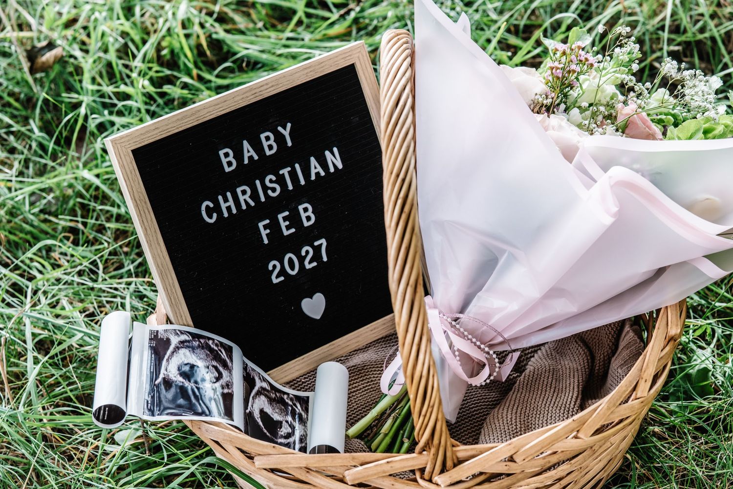 Baby Christian Feb 2027 pregnancy announcement letter board with ultrasound photos in a wicker basket on grass.