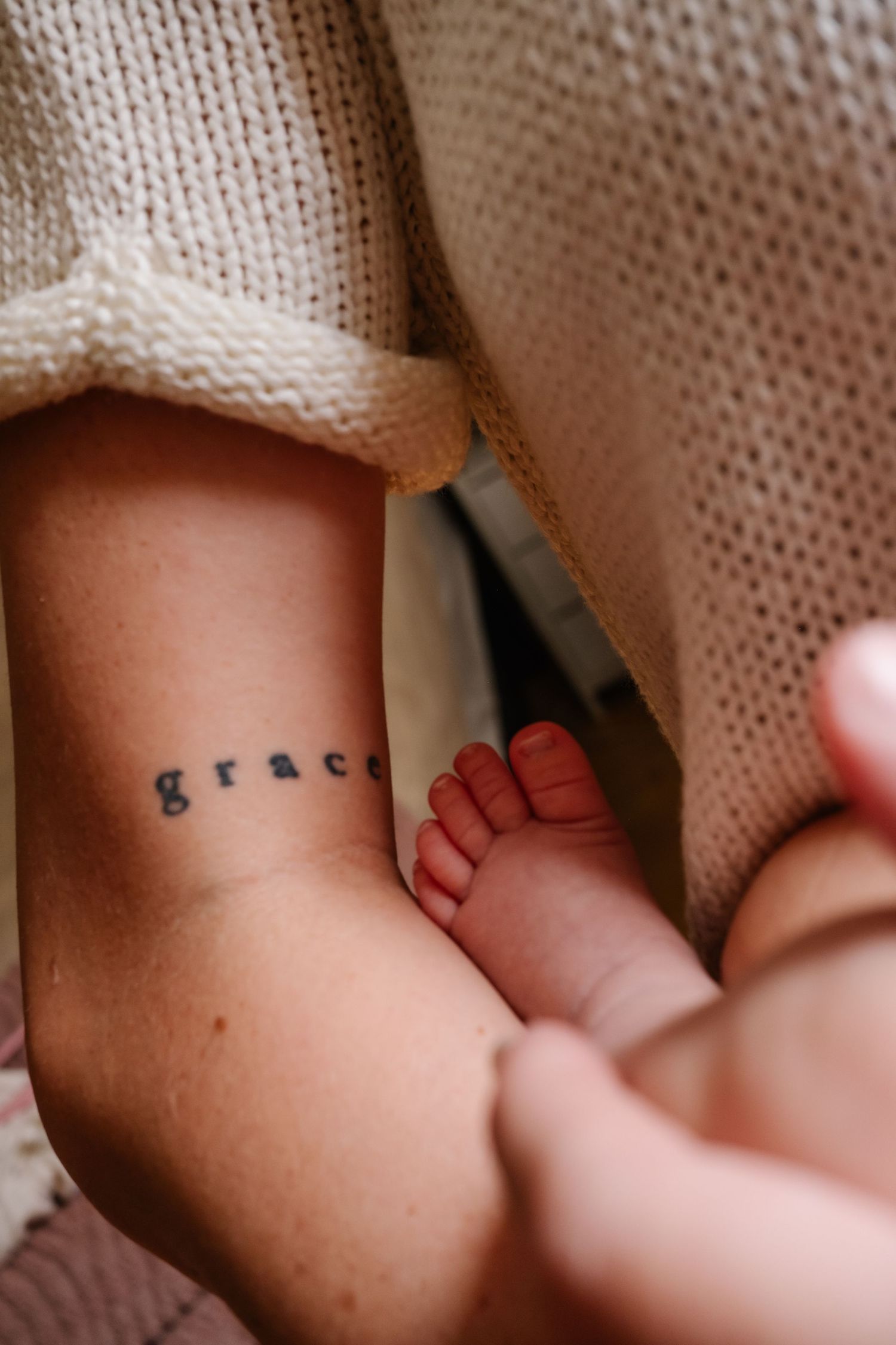 Close-up of an ankle tattoo reading 'grace' next to a newborn baby's tiny feet.