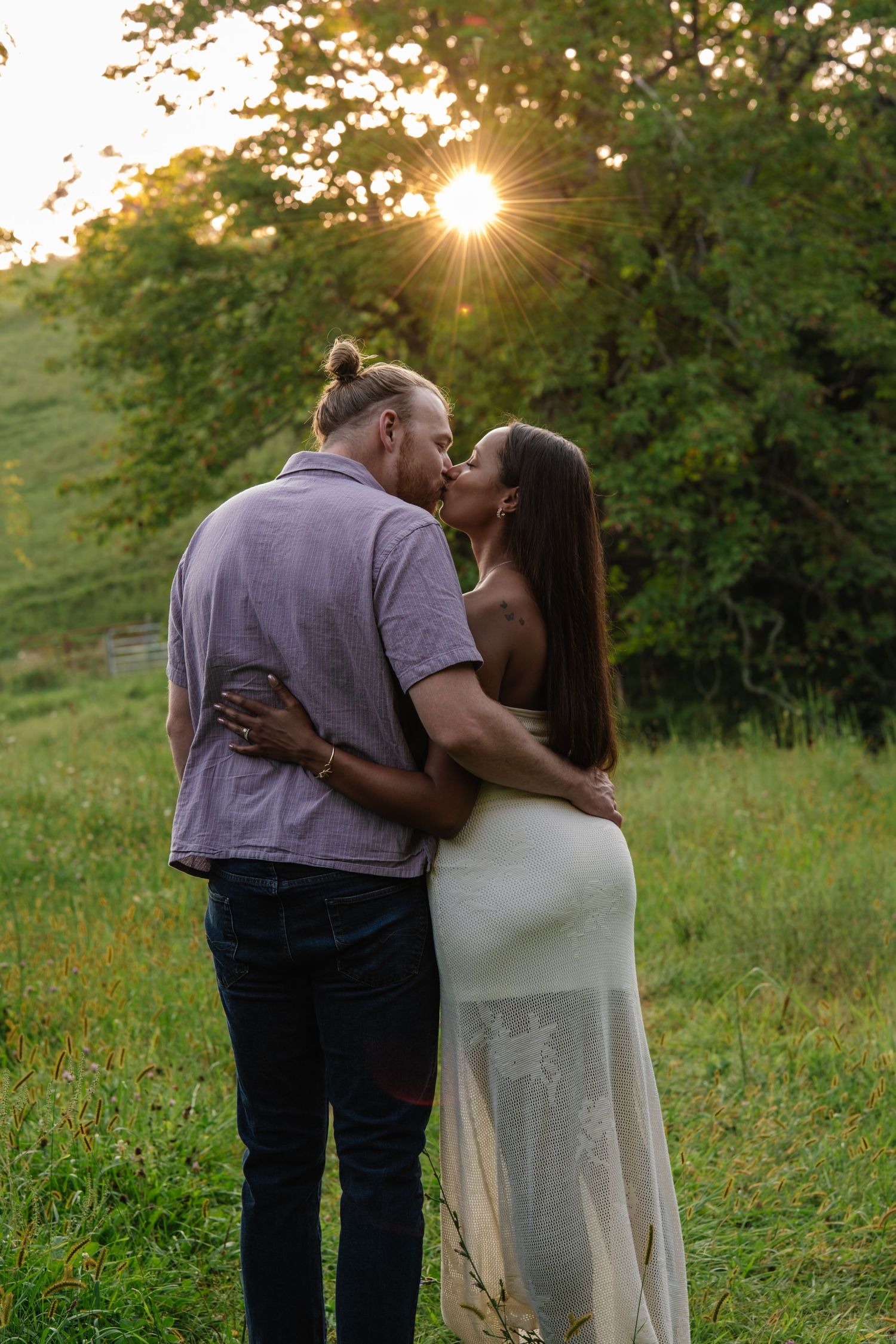 Couple sharing a romantic kiss at golden hour sunset in a lush green meadow, woman in white dress, man in casual attire.