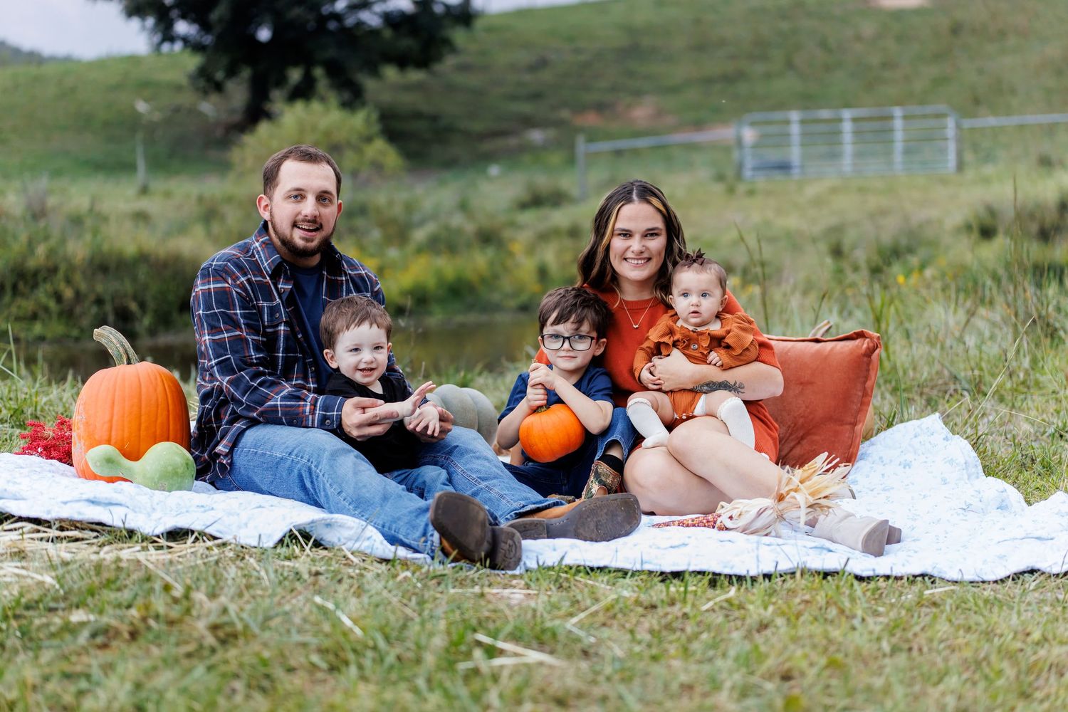 Family of five enjoying a fall picnic outdoors with pumpkins on a white blanket in a green field.