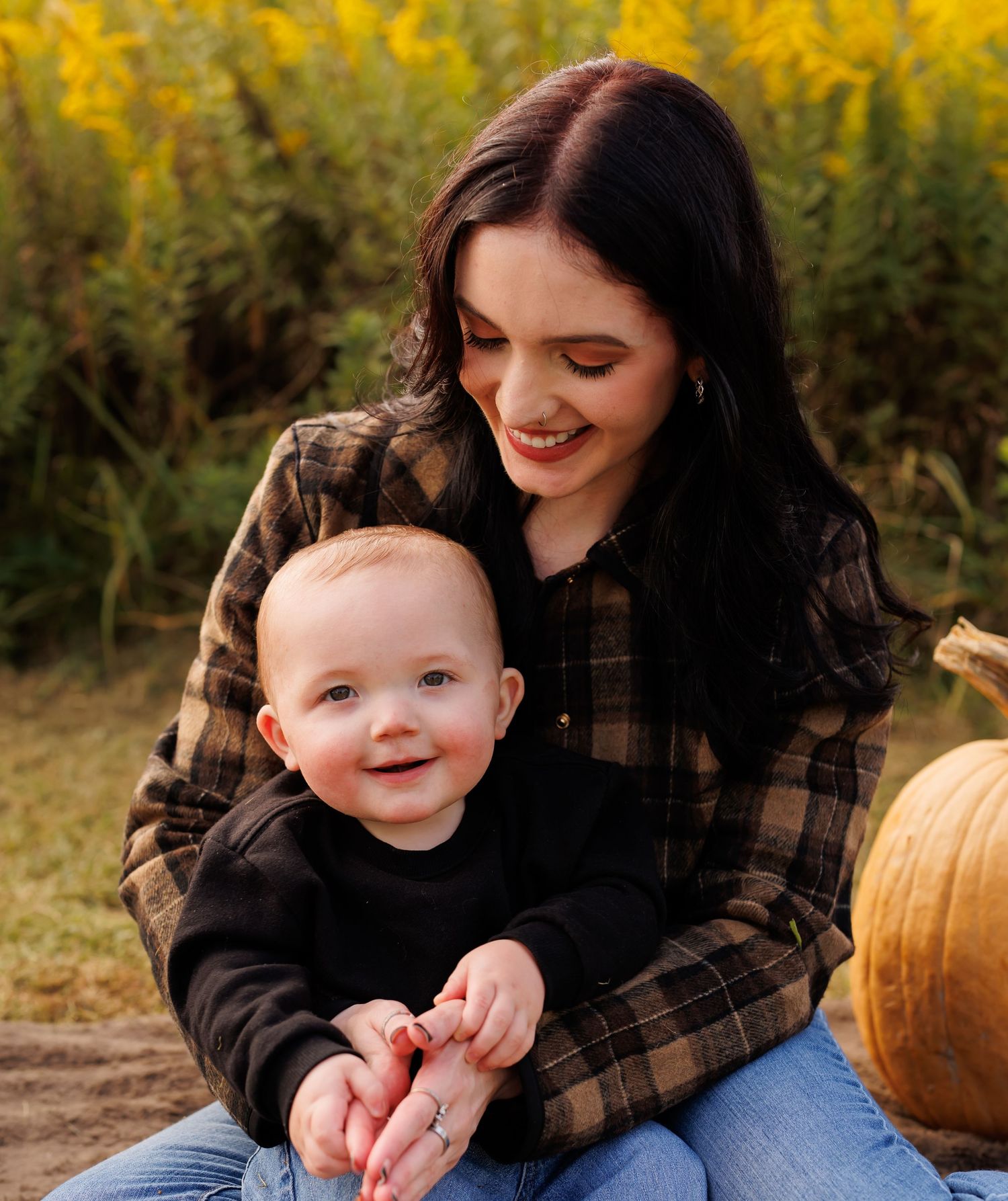 A smiling young woman with dark hair holds a happy baby outdoors near pumpkins and golden flowers in fall.