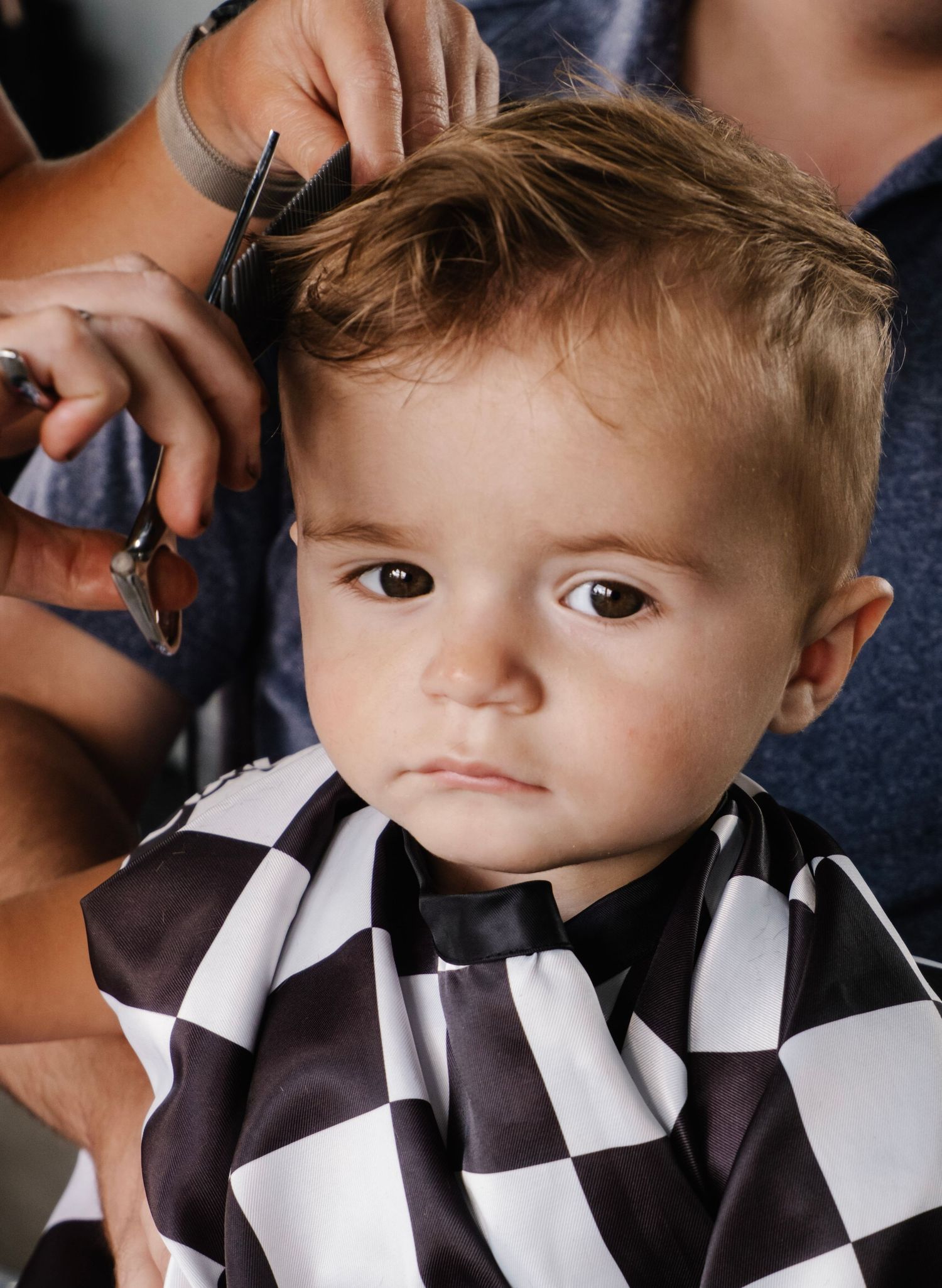 A blonde toddler boy in a checkered cape gets his first haircut with scissors at a salon.