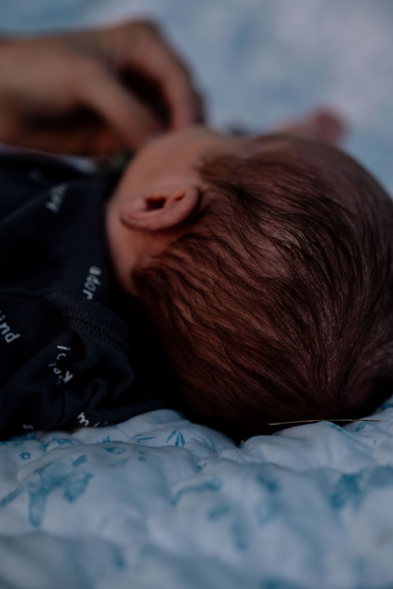 Close-up of a newborn baby with dark hair lying on a light blue blanket wearing a navy onesie.