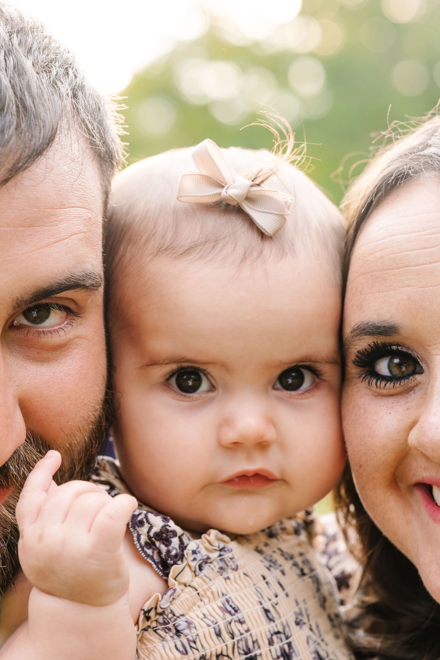 Parents and baby girl with bow headband pose cheek-to-cheek outdoors in a warm, bokeh-lit family portrait.