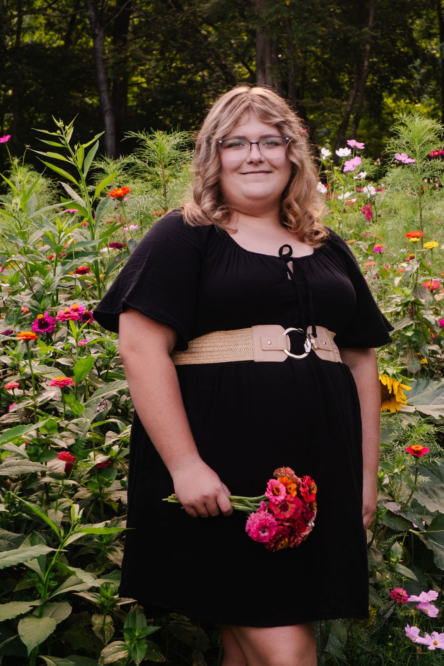 Smiling blonde woman in black dress holds zinnia bouquet in colorful flower garden.