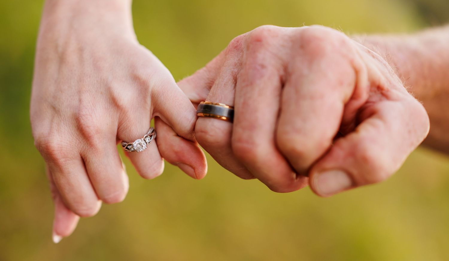 Two hands with wedding rings gently linking pinky fingers against a soft green bokeh background.