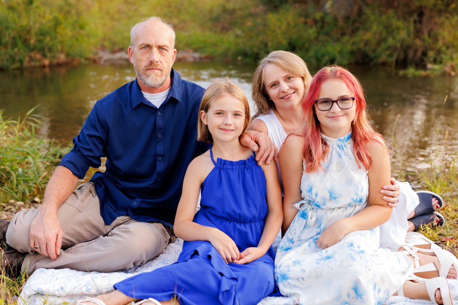 Family of four smiling outdoors near a creek, sitting on a blanket in a sunny natural setting.
