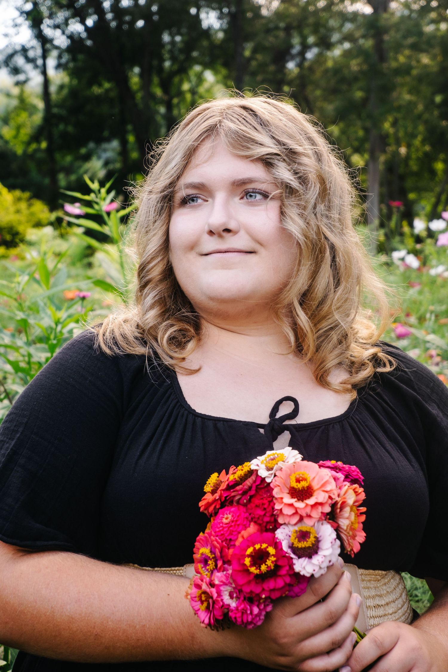 Smiling blonde woman holding vibrant pink and orange zinnia bouquet in garden.