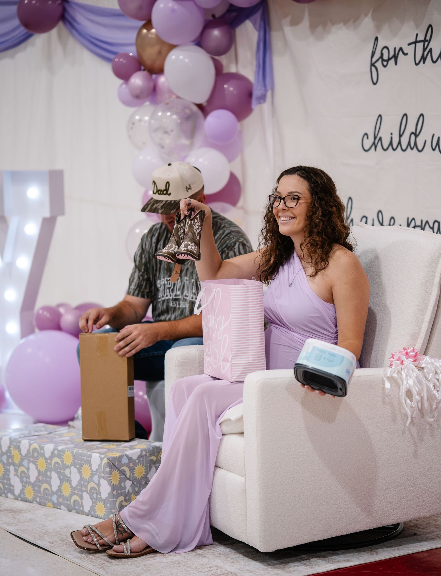Woman in lavender outfit smiles at baby shower with purple balloon arch and gift boxes.