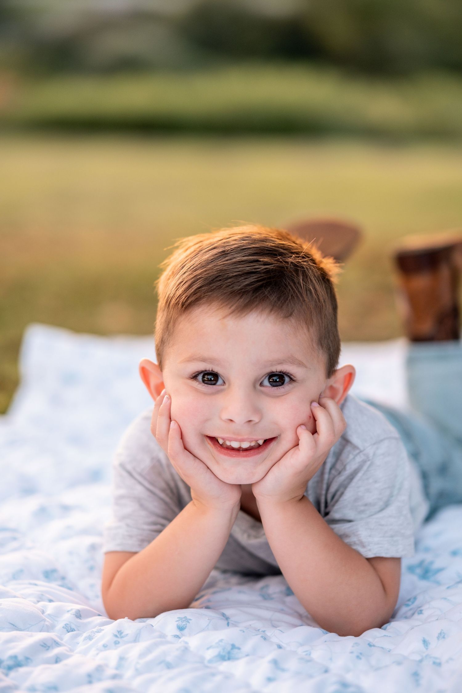 Young boy smiling with hands on cheeks lying on a blue floral blanket outdoors in a sunny field.