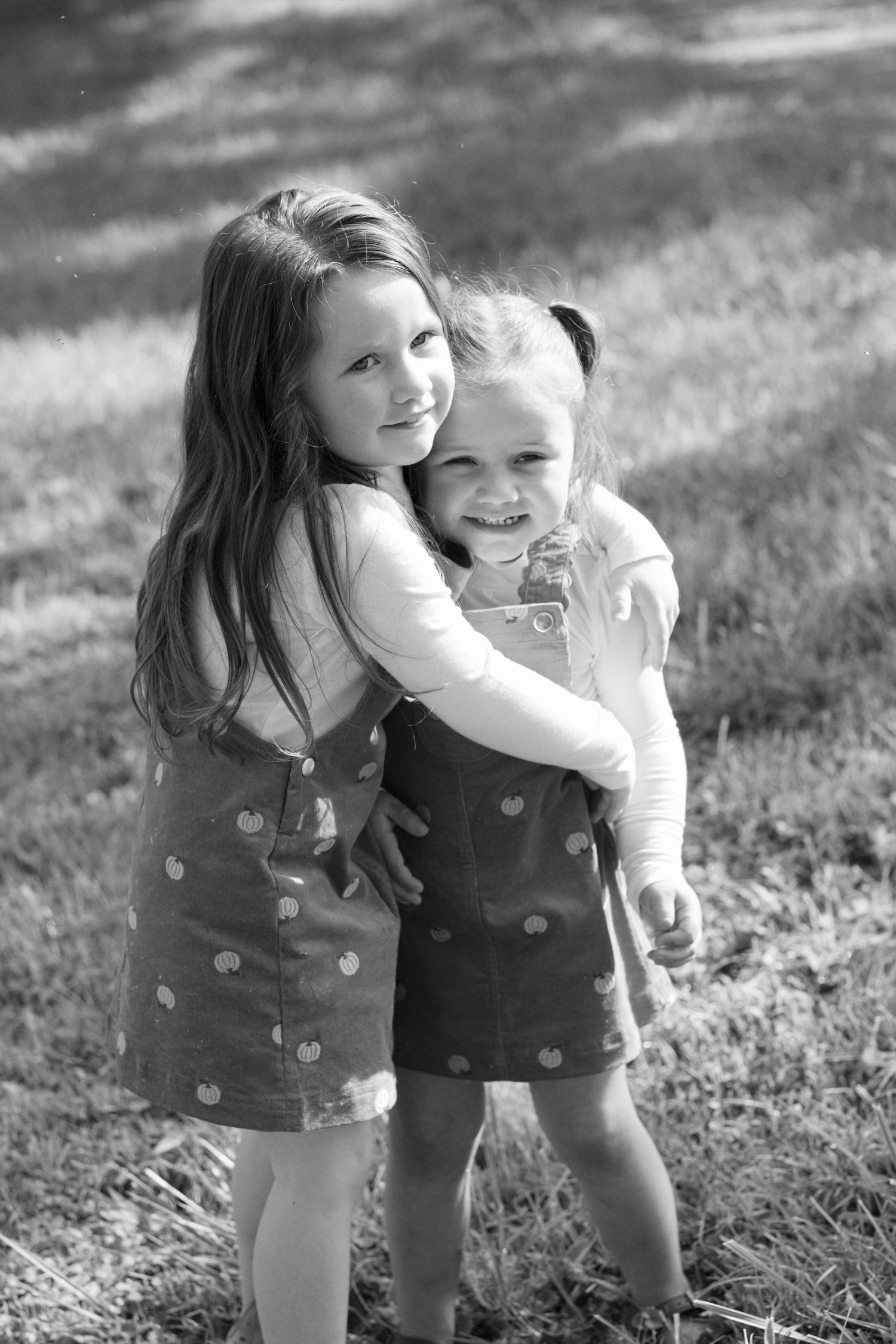 Two young sisters in matching polka dot dresses share a warm hug outdoors in this black and white photo.