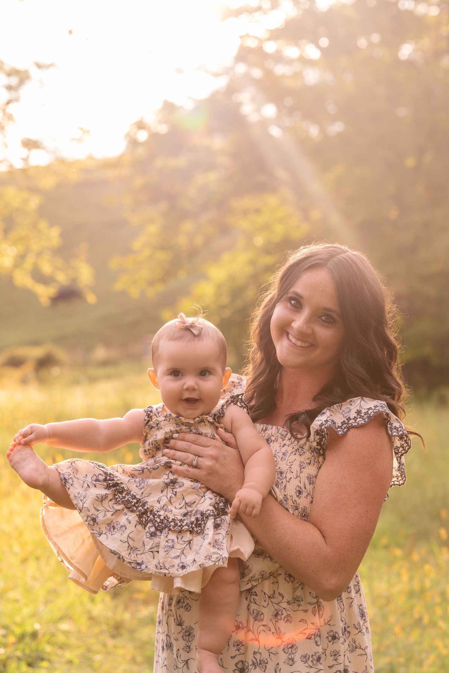 A smiling mother holds her baby girl in matching floral dresses outdoors in golden sunlight.