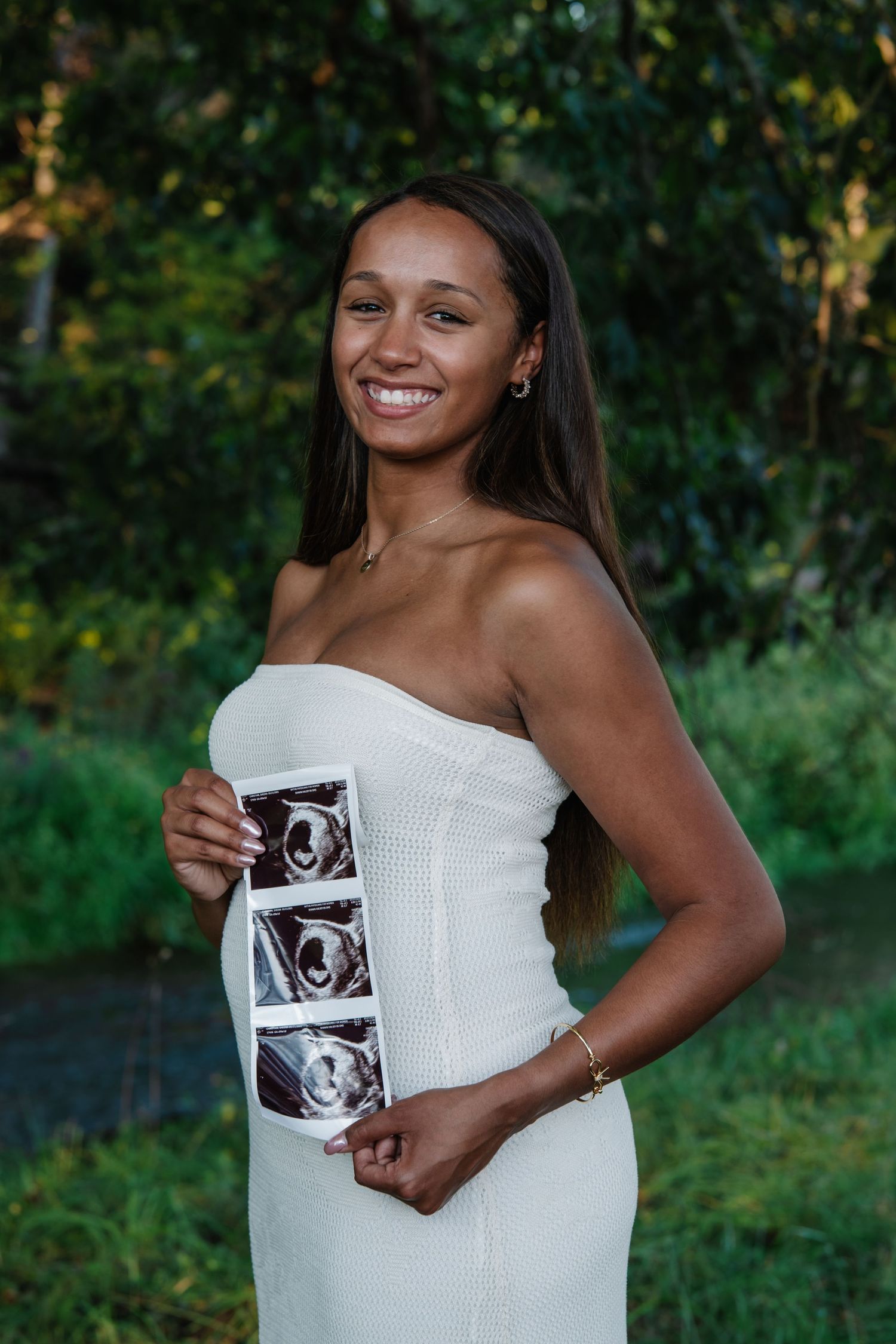 Smiling pregnant woman in white strapless dress holding ultrasound photos outdoors with green trees in background.