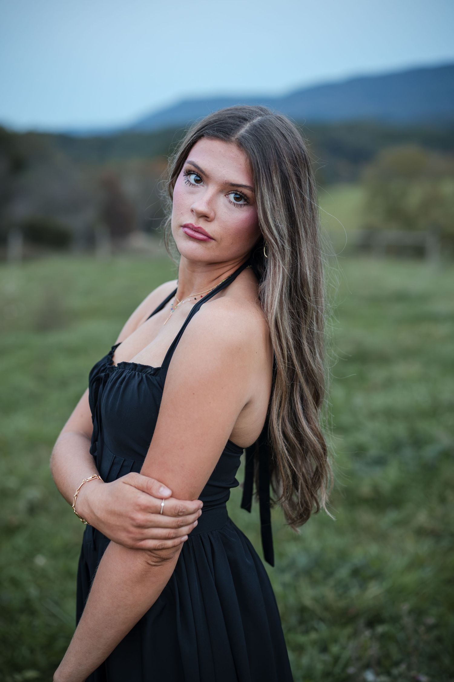 Young woman in black sundress poses outdoors with mountain backdrop, long brown hair, gold jewelry.