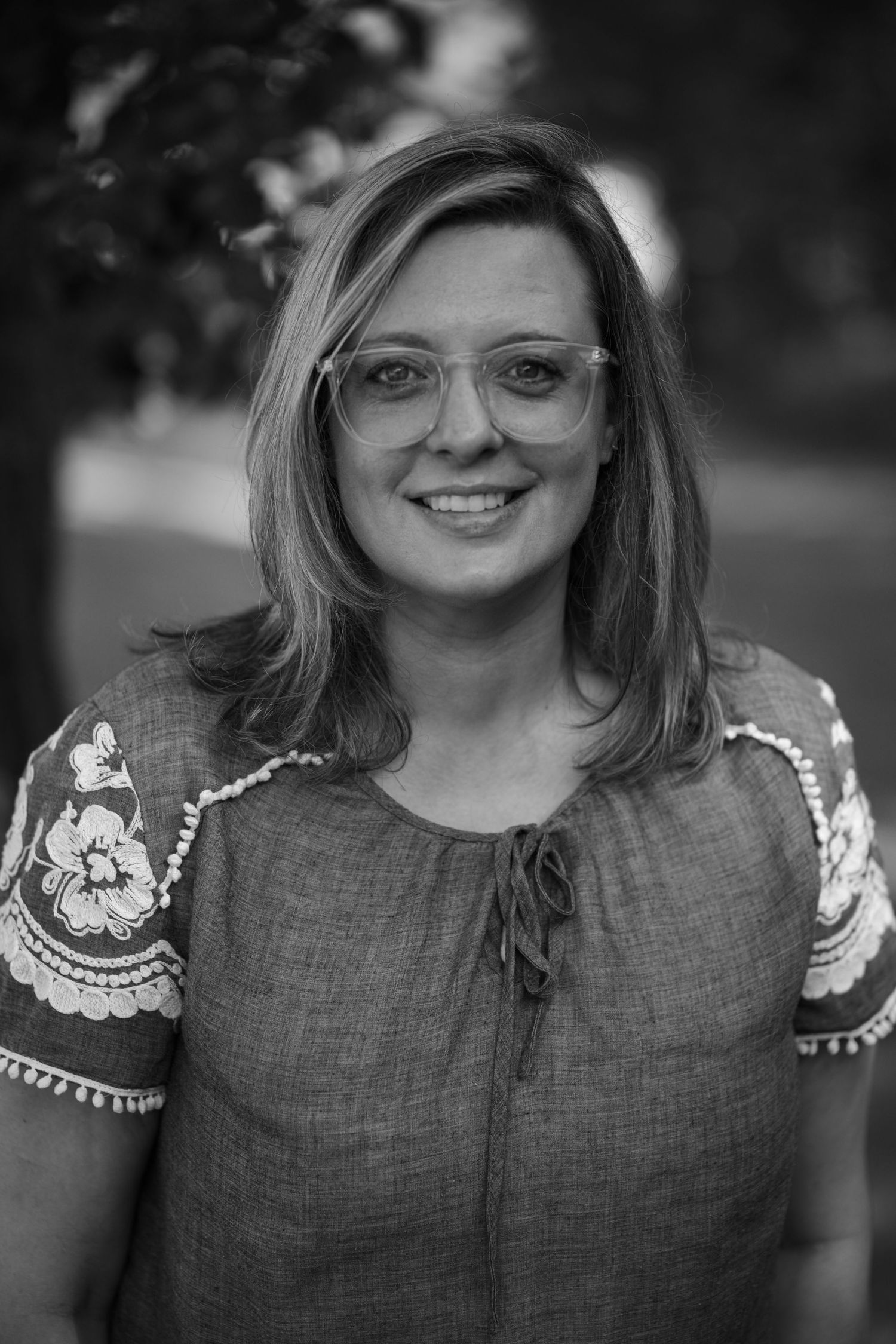 Smiling woman with glasses and shoulder-length hair wearing an embroidered blouse, black and white outdoor portrait.