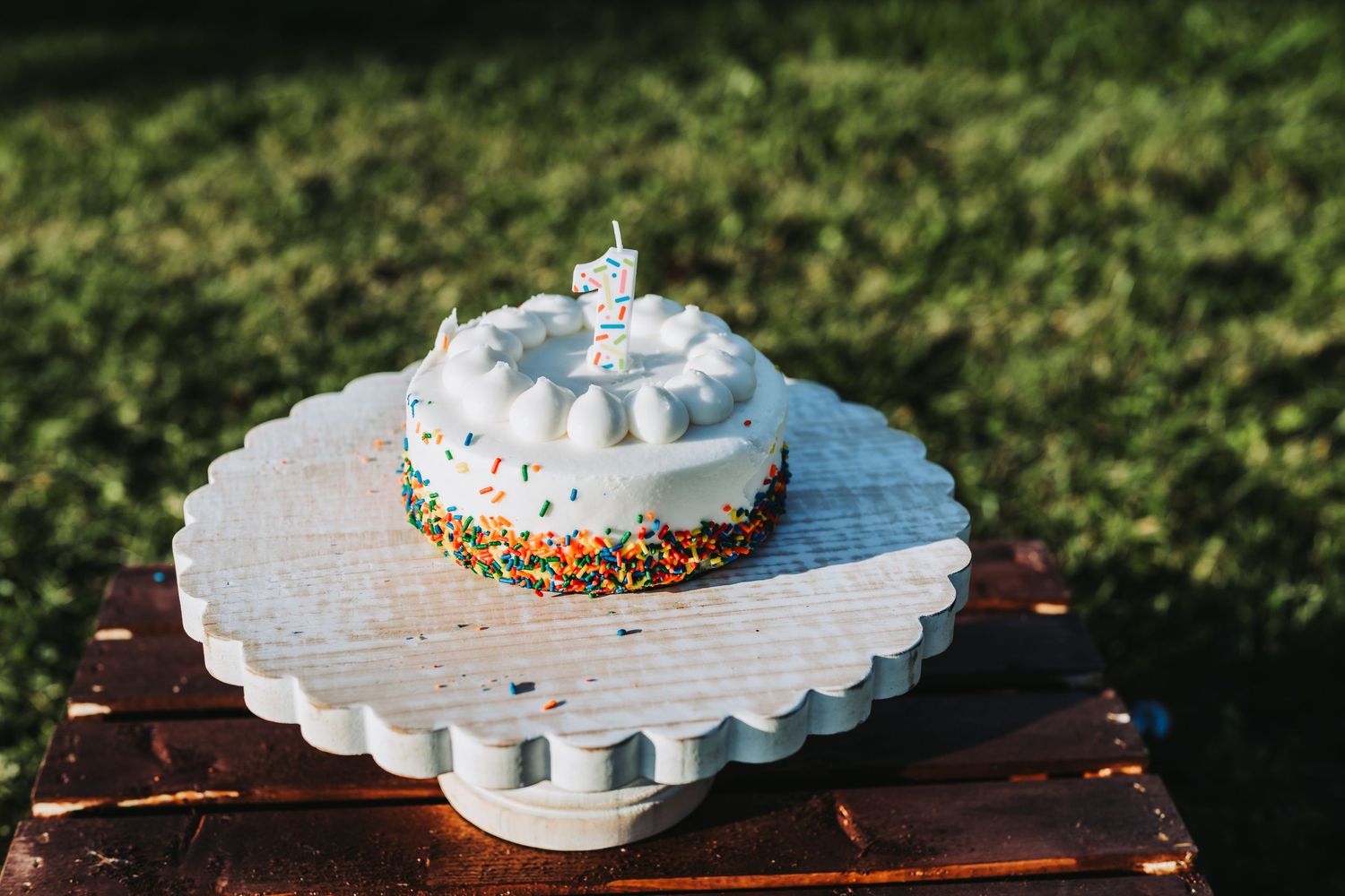 A small white birthday cake with colorful sprinkles and a candle on a decorative stand outdoors.