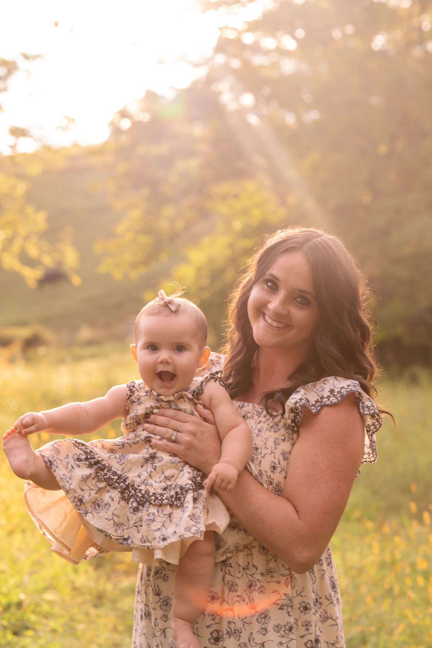 A smiling woman holds a happy baby girl in matching floral dresses during a golden hour outdoor photo session.