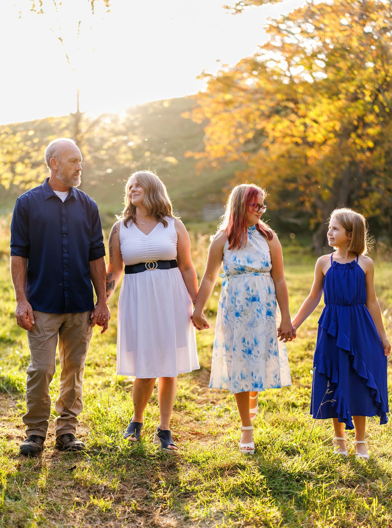 Family of four holding hands in a sunlit field with autumn trees, wearing elegant dresses and casual attire.