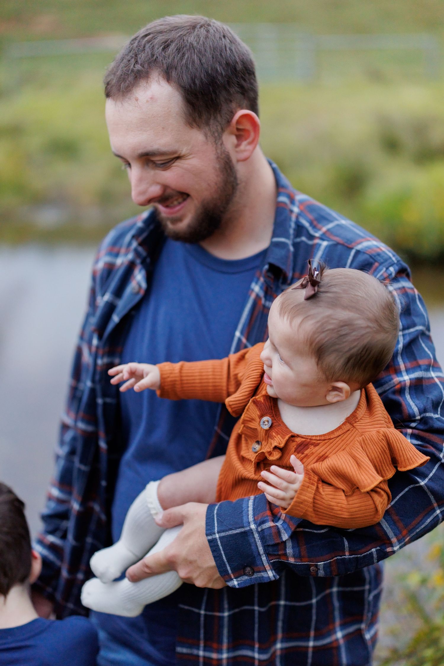 A smiling bearded man in a plaid shirt holds a baby girl in an orange outfit outdoors near a pond.