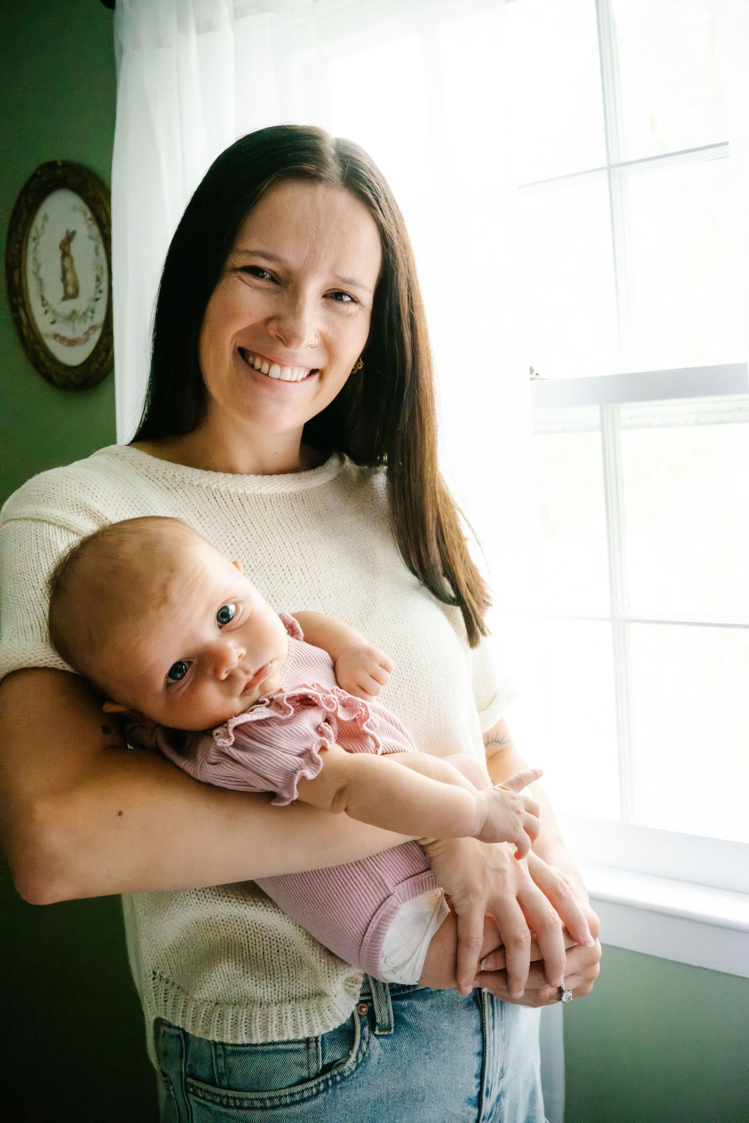 Smiling brunette woman holding a newborn baby in pink outfit near a bright window indoors.