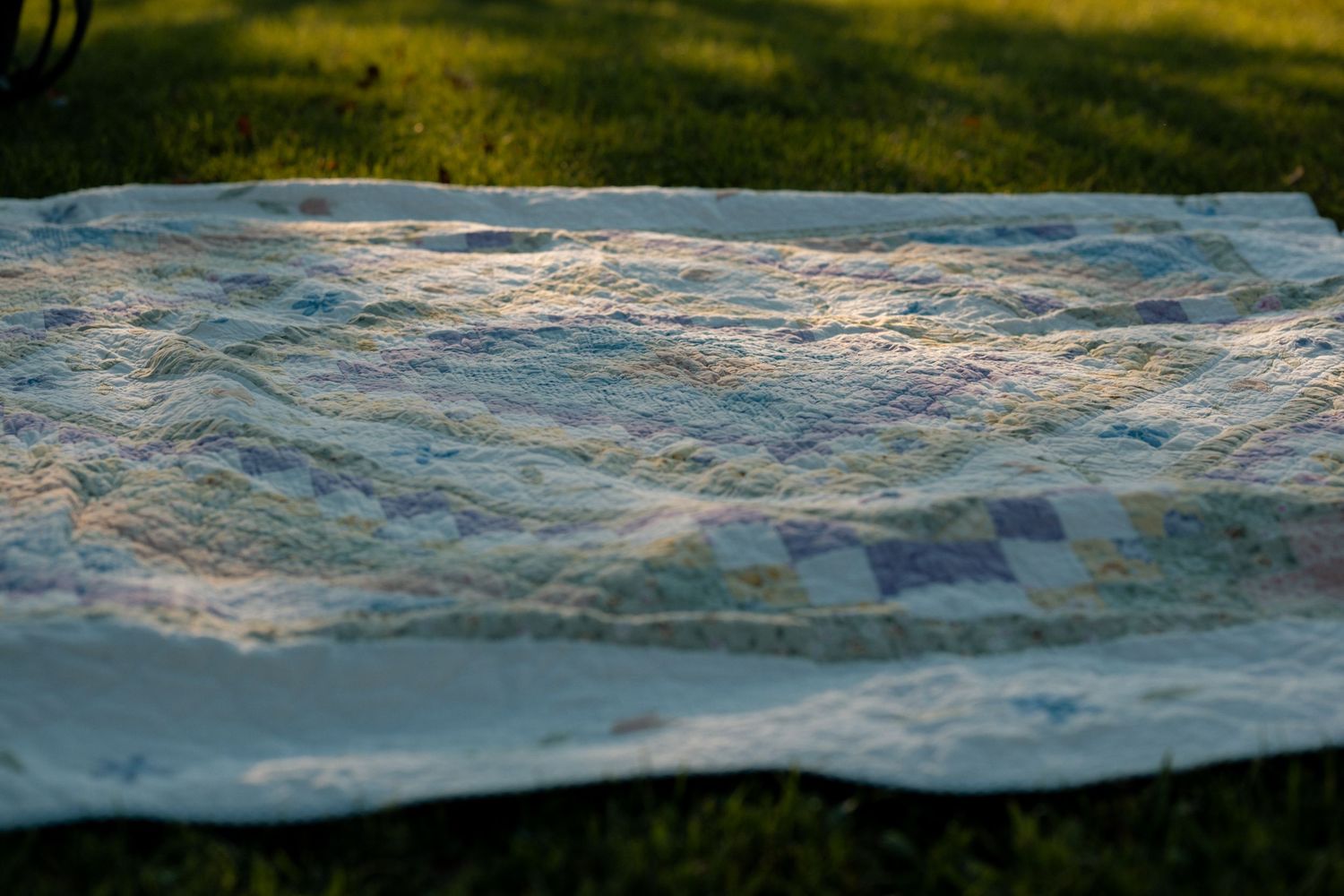 A patchwork quilt with blue and green squares laid flat on green grass outdoors in soft natural light.