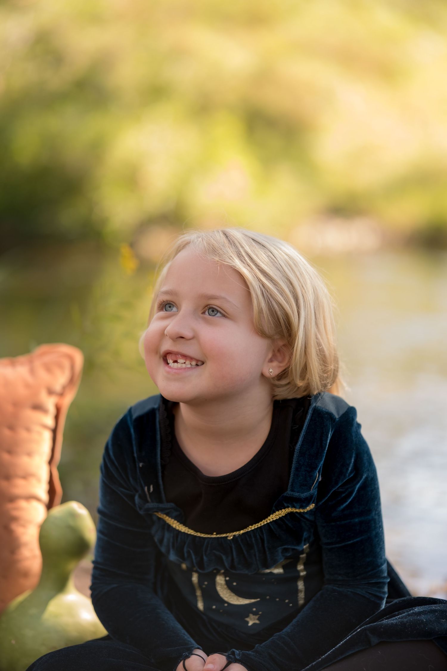 Smiling blonde child in navy velvet outfit sitting outdoors with bokeh green background during golden hour.