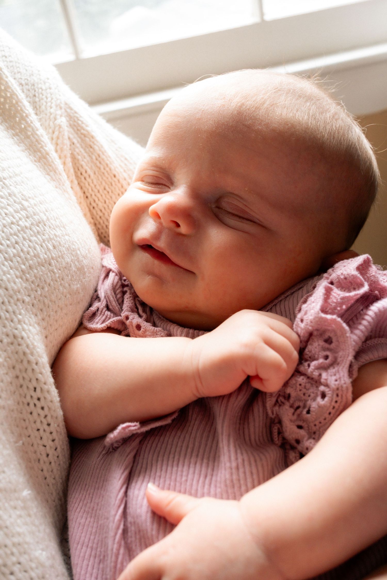A peaceful newborn baby smiles while sleeping, dressed in a pink outfit, cradled in loving arms near a bright window.