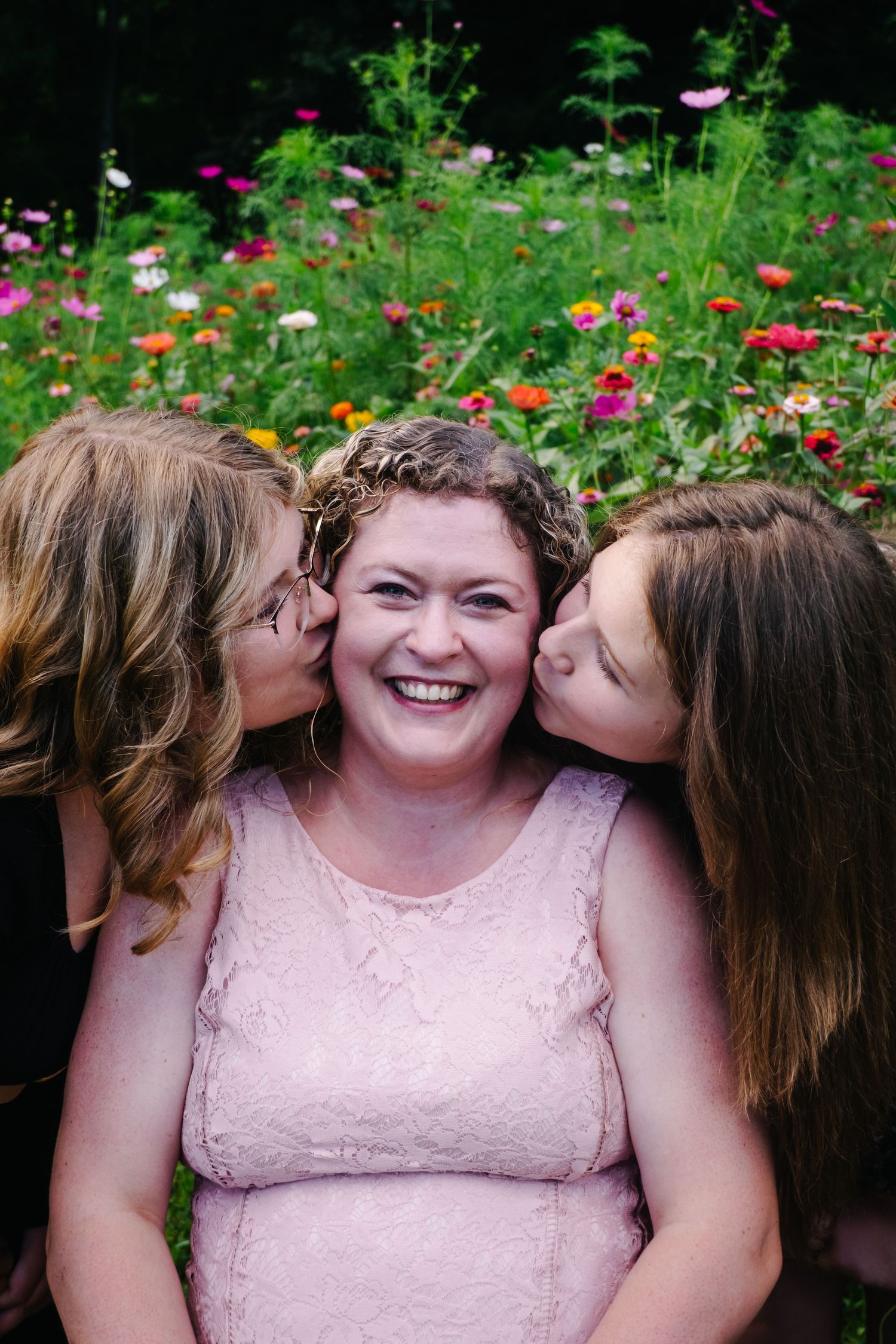 Woman in pink dress laughing as two girls kiss her cheeks in a colorful flower garden.