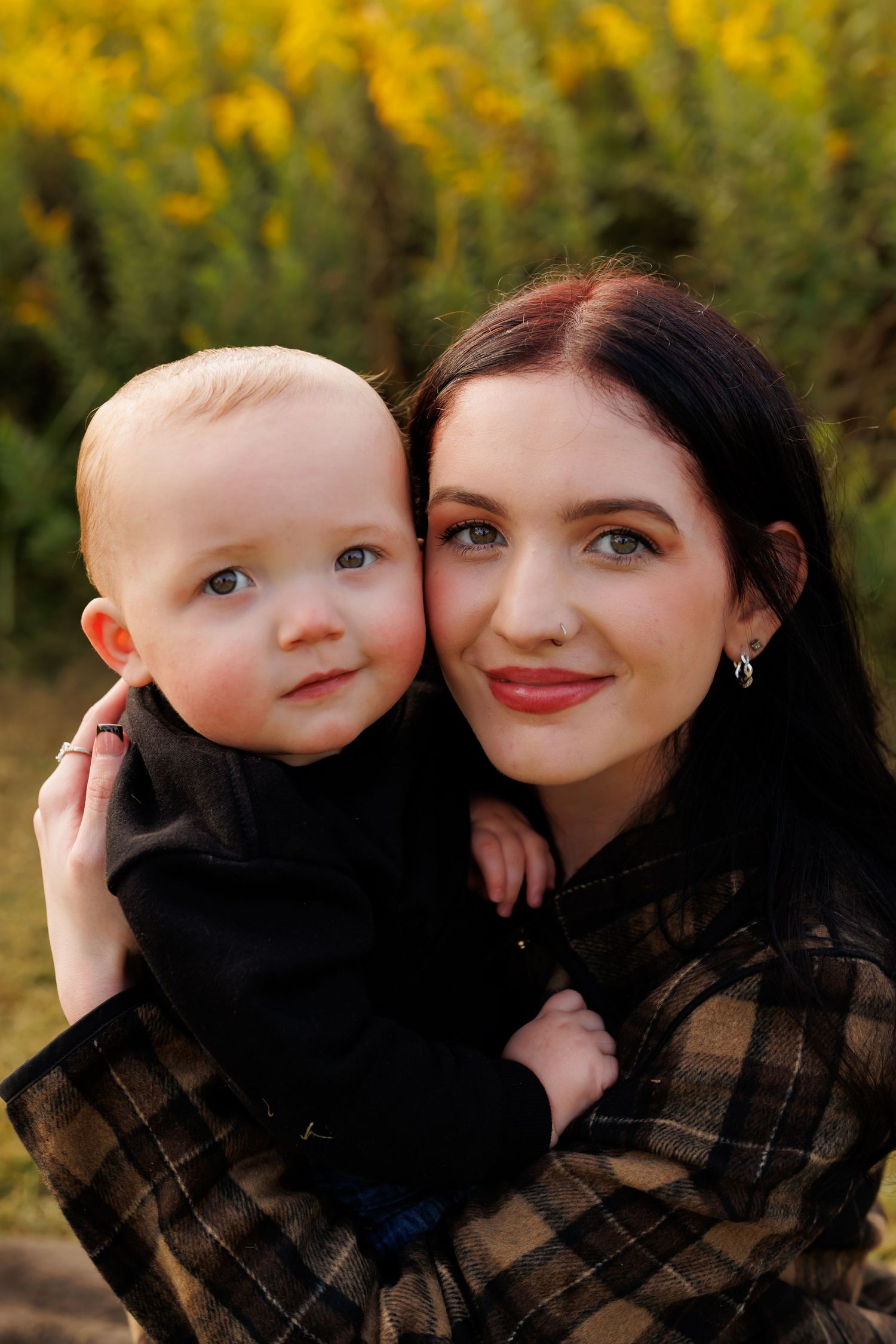 Young mother with dark hair holds smiling baby boy outdoors near yellow wildflowers in autumn.