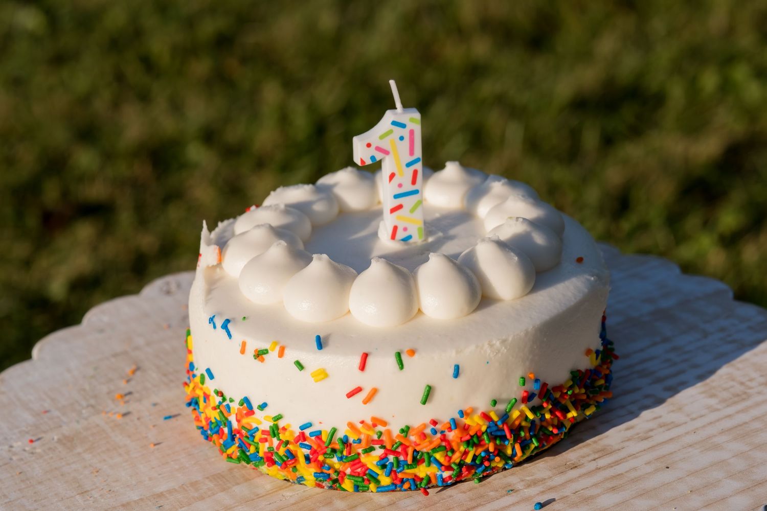 White frosted first birthday cake with colorful sprinkles and a number 1 candle on a wooden surface outdoors.