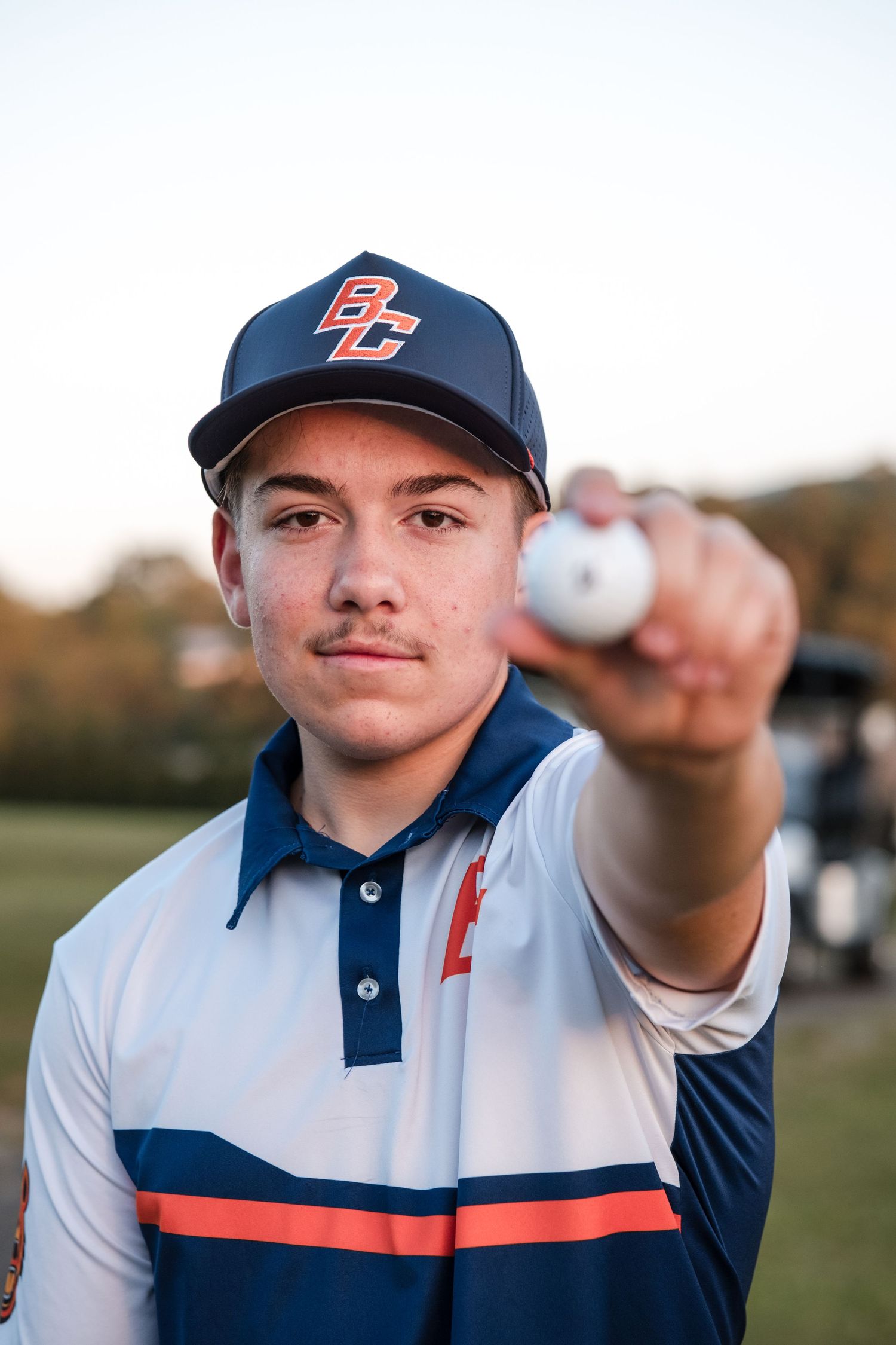 Young golfer in BC cap holds golf ball toward camera on a green course at golden hour.