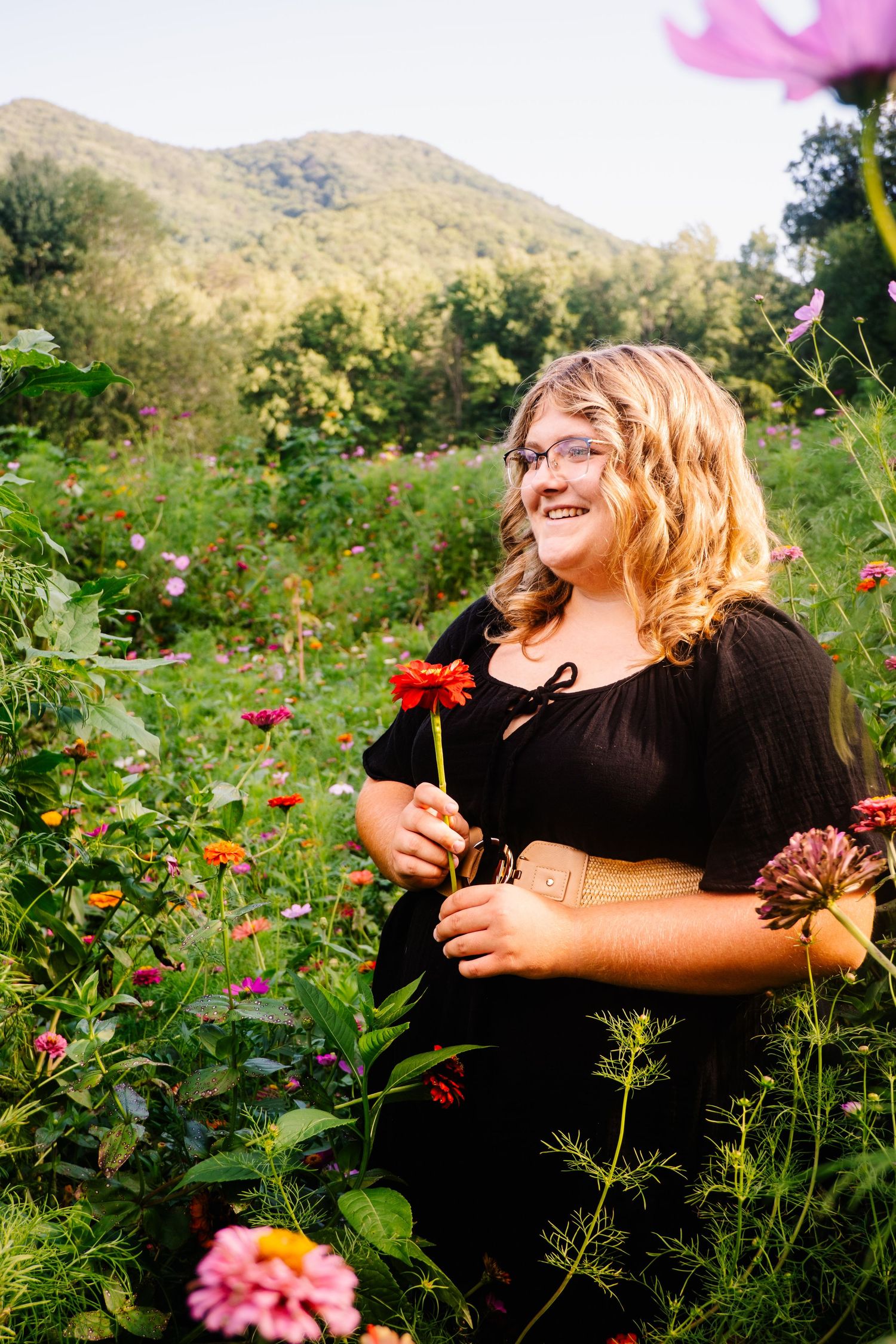 A smiling blonde teen in a black dress holds a red zinnia in a colorful mountain flower field.