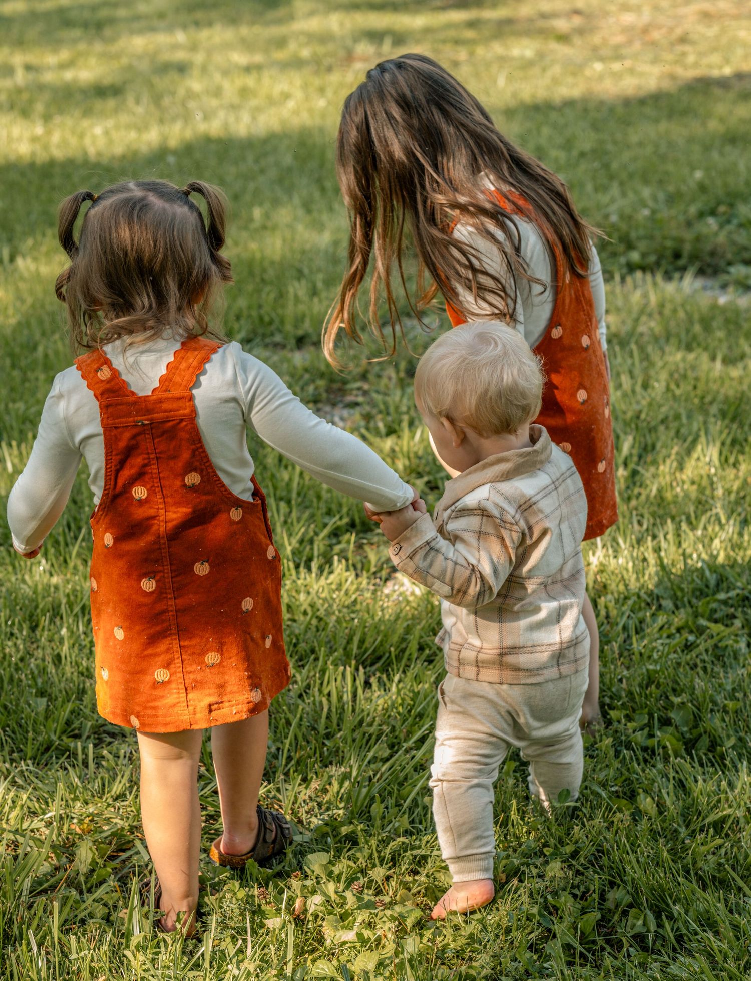 Three young children holding hands on green grass, two girls in orange dresses and a toddler boy in plaid.