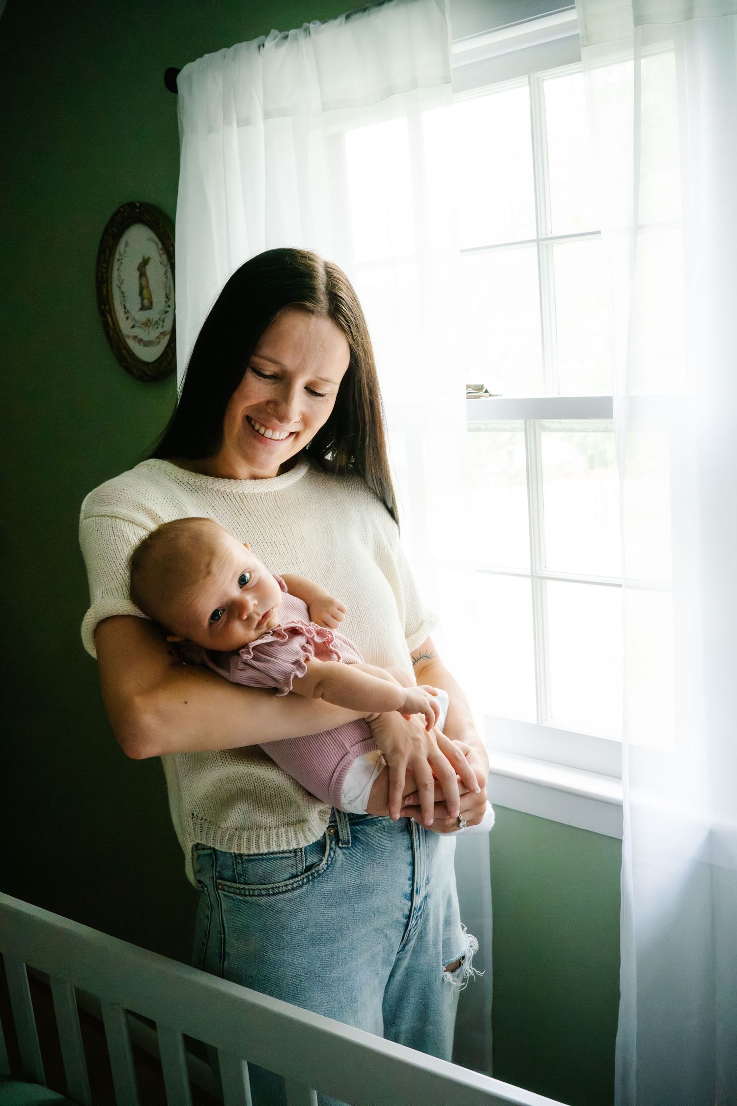 Smiling mother holding newborn baby in purple outfit near window in green nursery room.