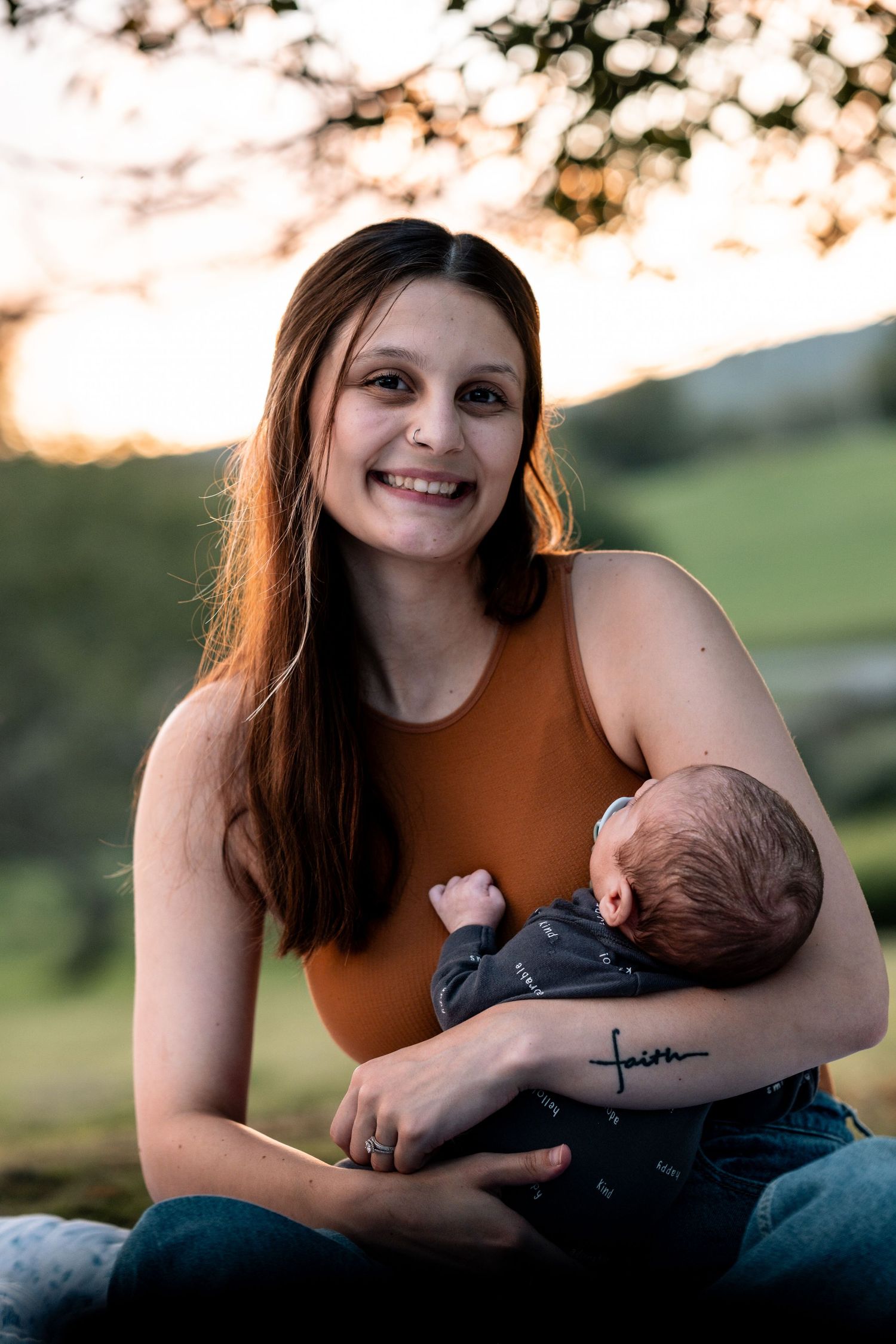Smiling young mother holding newborn baby outdoors at golden hour with faith tattoo visible.