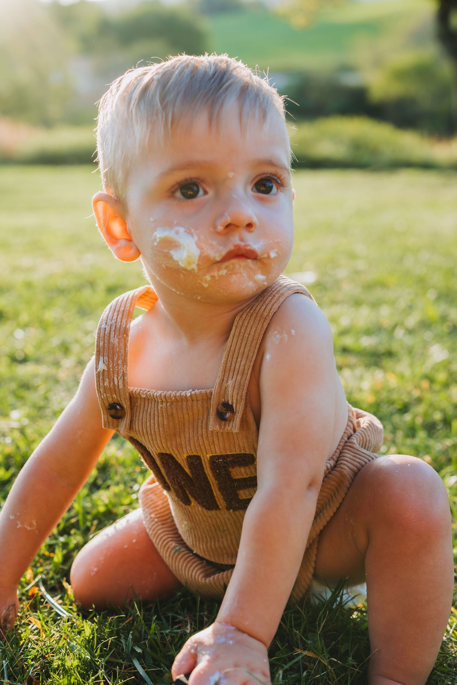 Messy toddler boy with cake on his face sitting on green grass outdoors in warm sunlight.