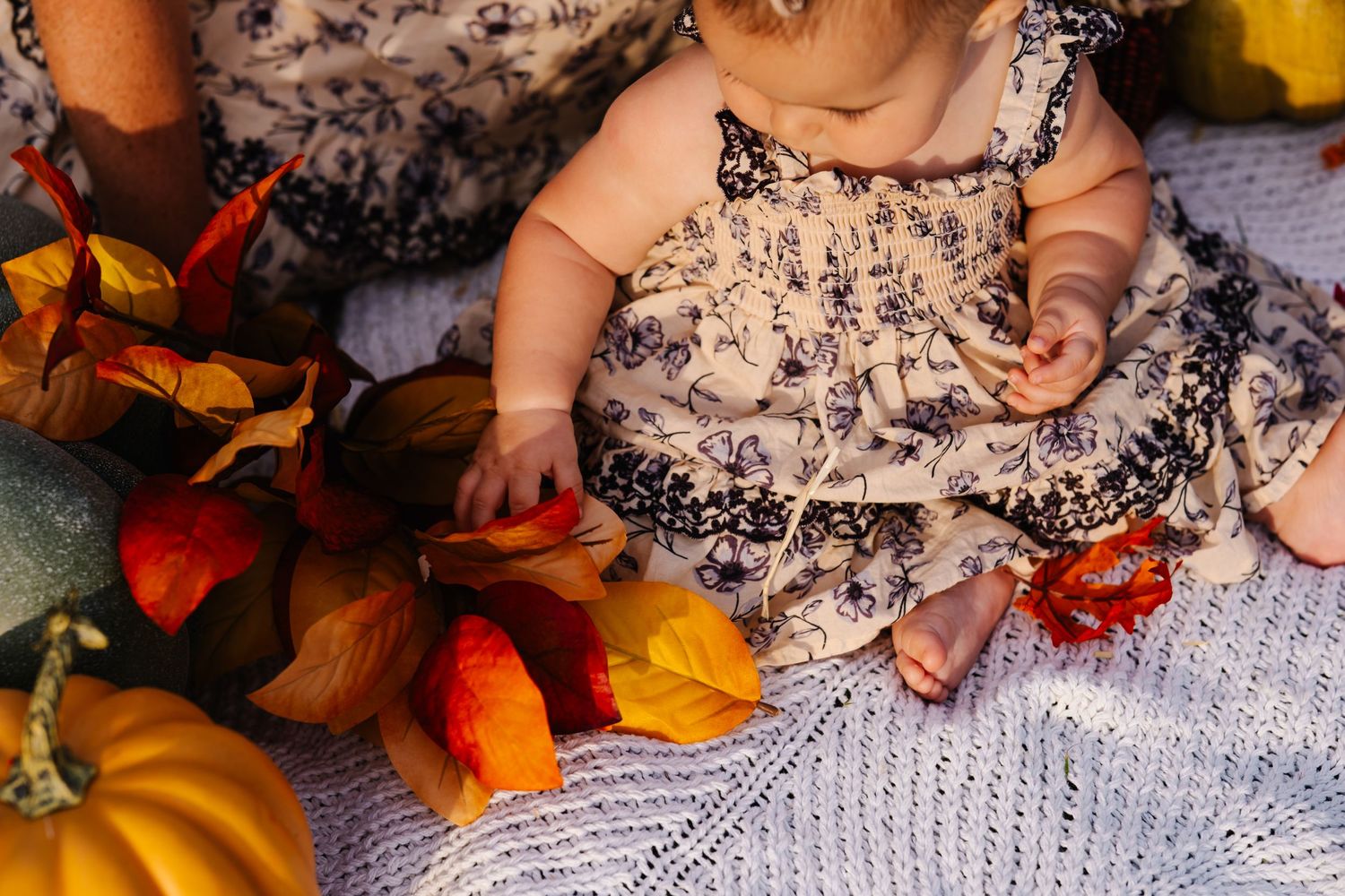 Baby in floral dress sits on knit blanket surrounded by autumn leaves and pumpkins in a fall-themed photo.