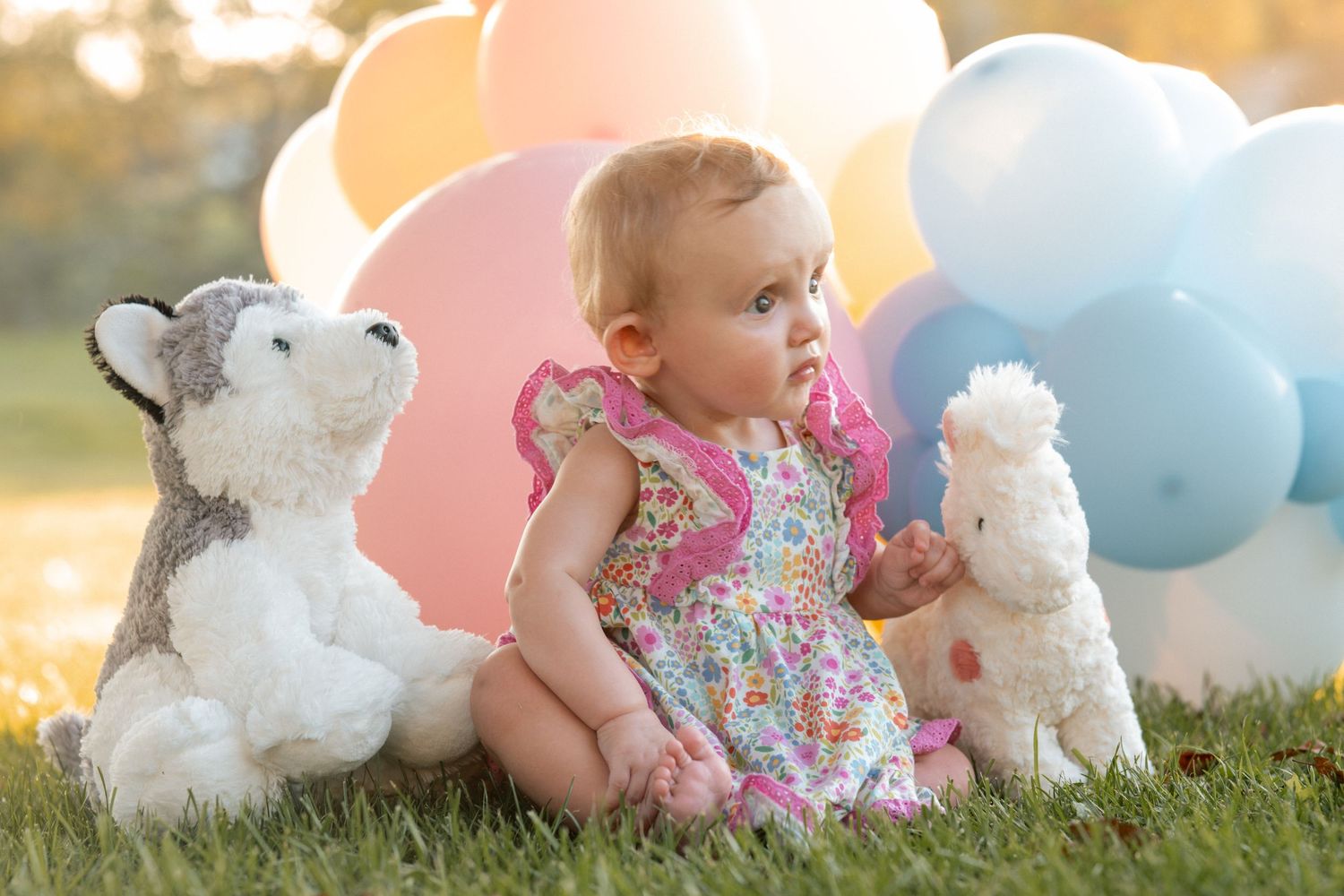Baby girl in floral dress sits on grass with stuffed animals and colorful balloons outdoors.