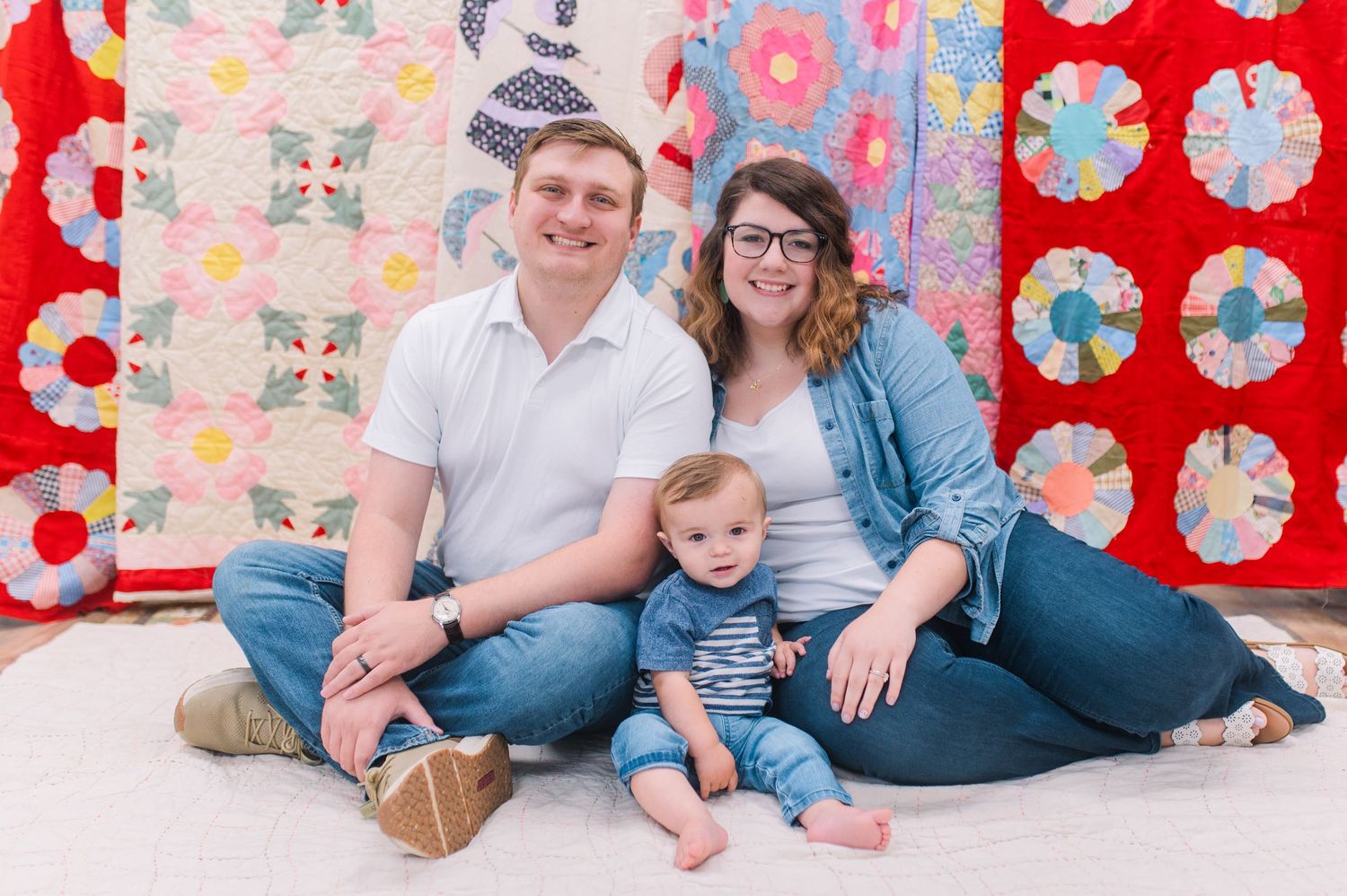 Young family with baby sits on floor in front of colorful vintage quilts for portrait photo.
