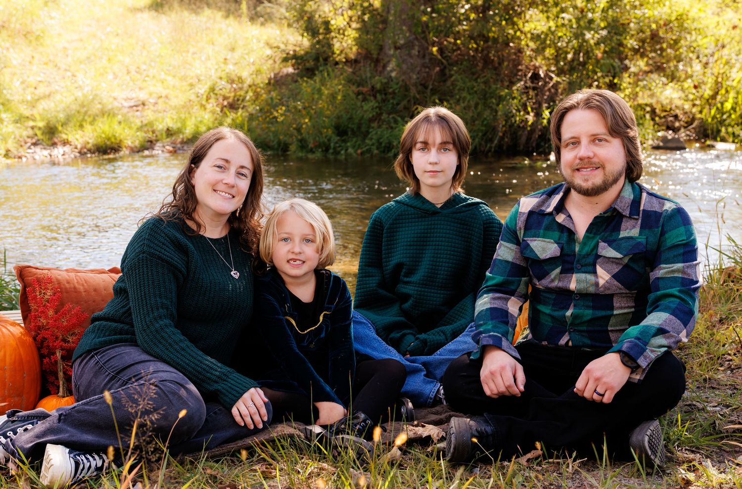 Family of four smiling together outdoors by a river, sitting on grass surrounded by autumn foliage.