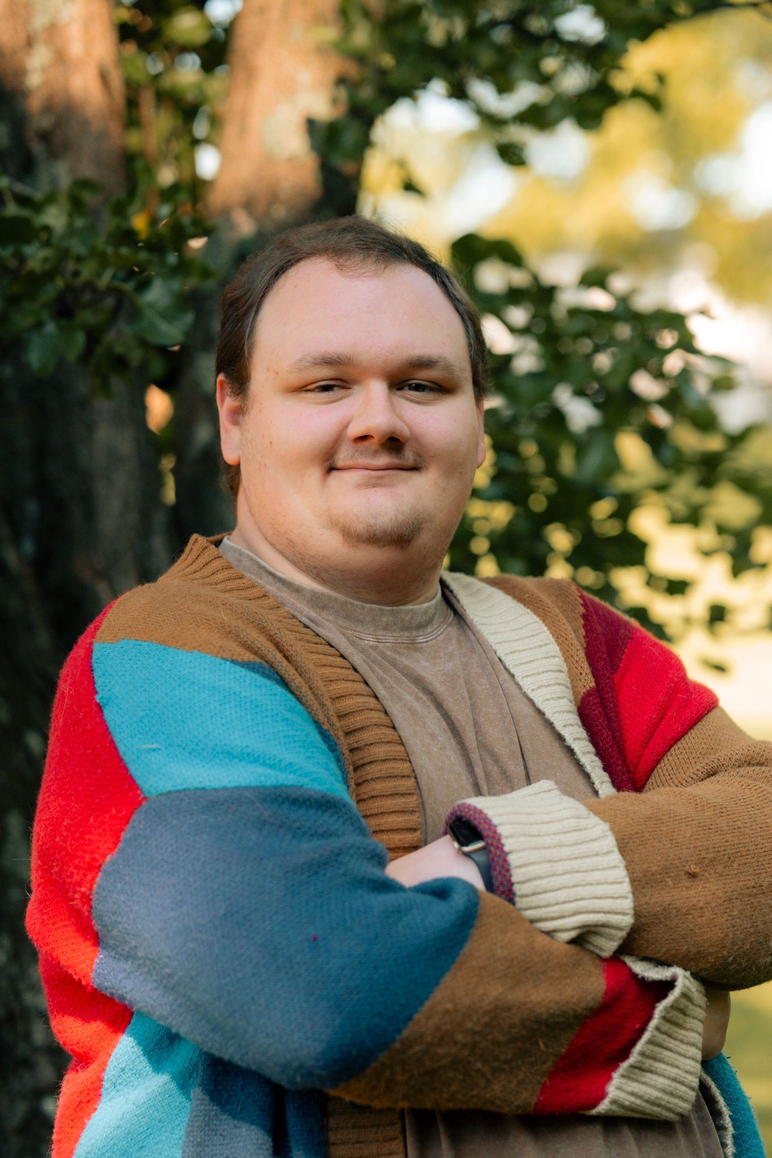 Young man in colorful striped cardigan stands arms crossed outdoors near trees, smiling confidently.