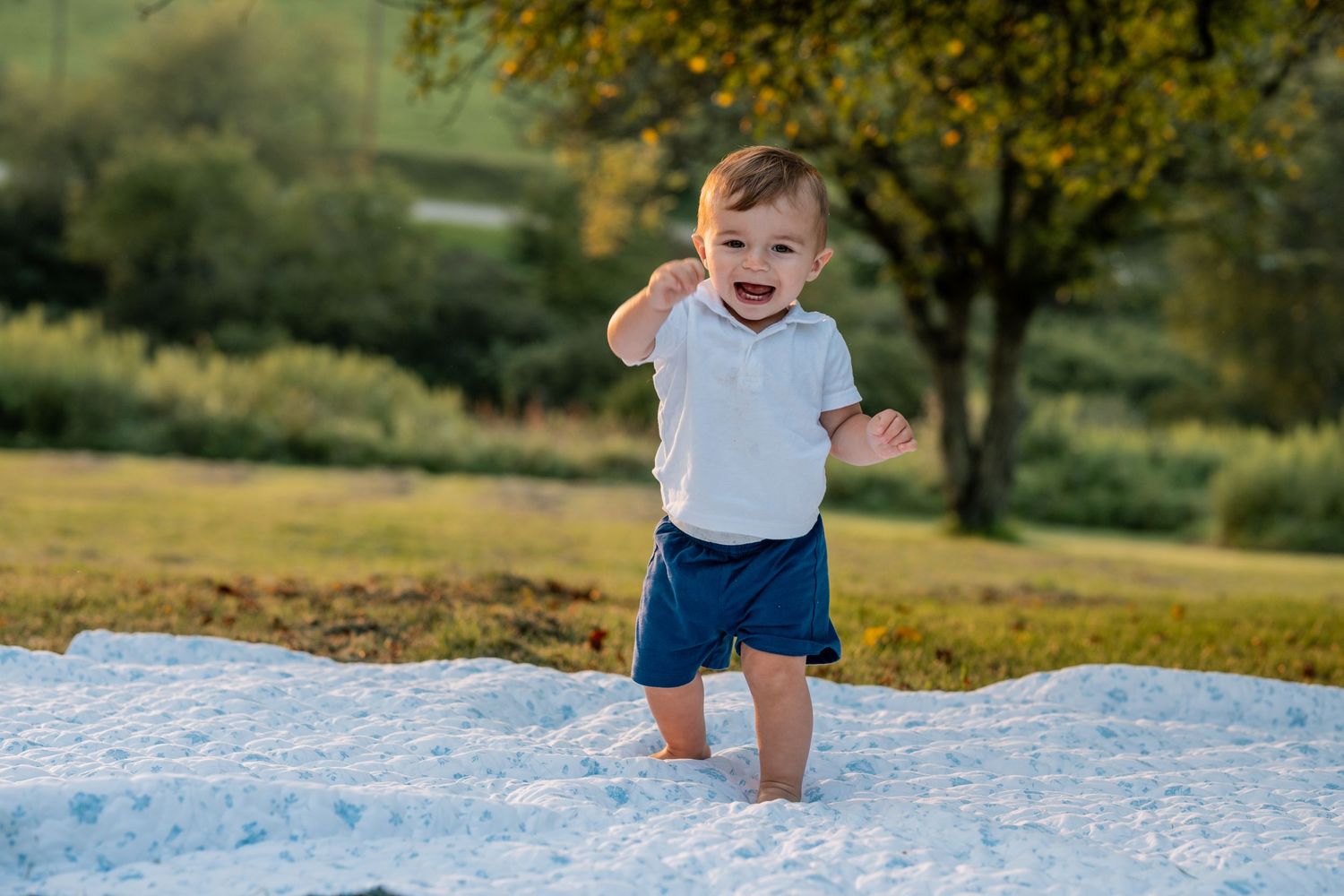 Happy toddler boy in white shirt and navy shorts playing on a white blanket in a green field.