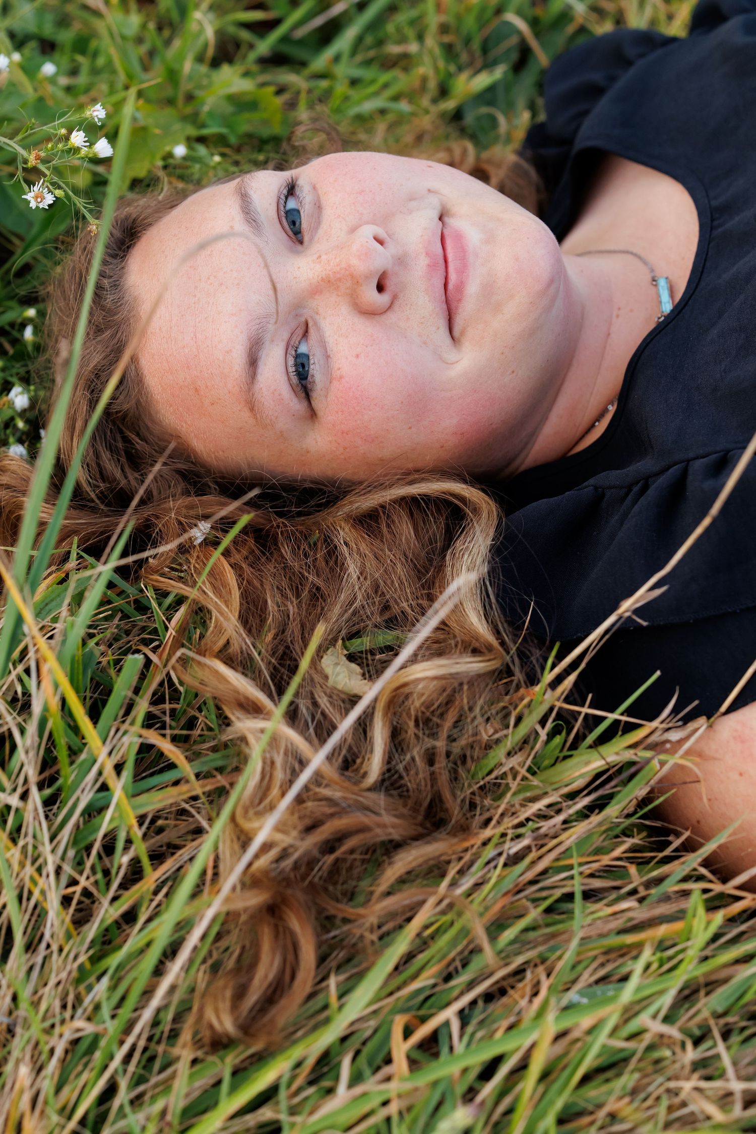 Young woman with long wavy hair lying in green grass field, looking up peacefully on a sunny day.