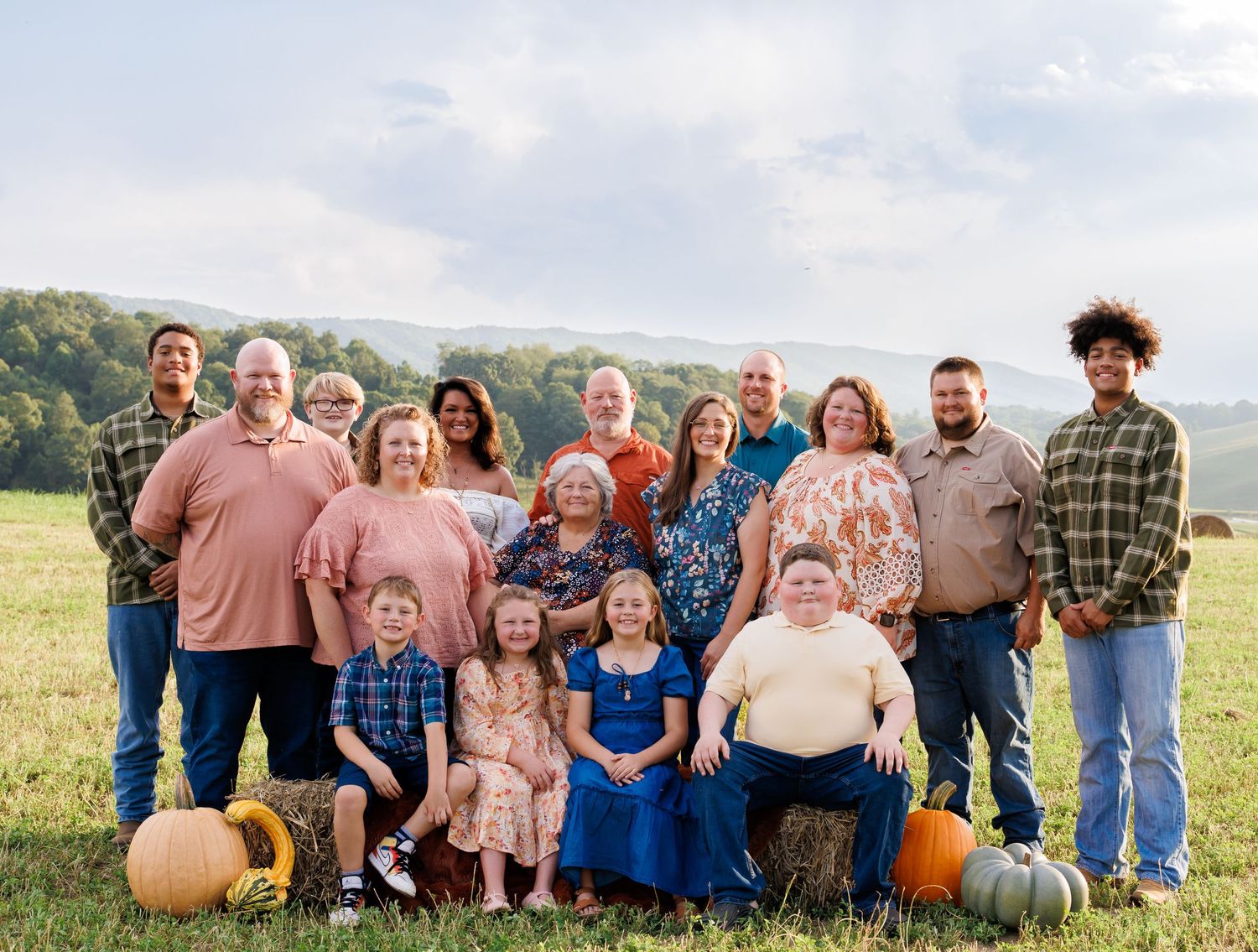 Large family group portrait outdoors in a green field with pumpkins, rolling hills, and a cloudy sky backdrop.