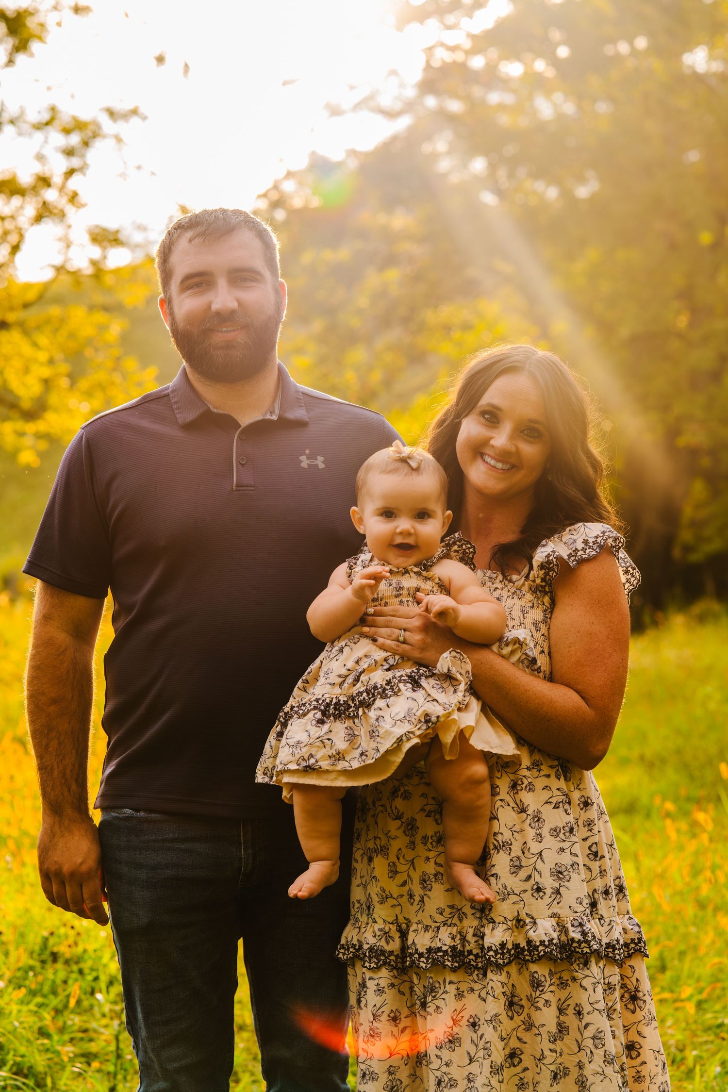A happy family of three poses outdoors in golden sunlight, with parents smiling and holding their baby girl in a floral dress.