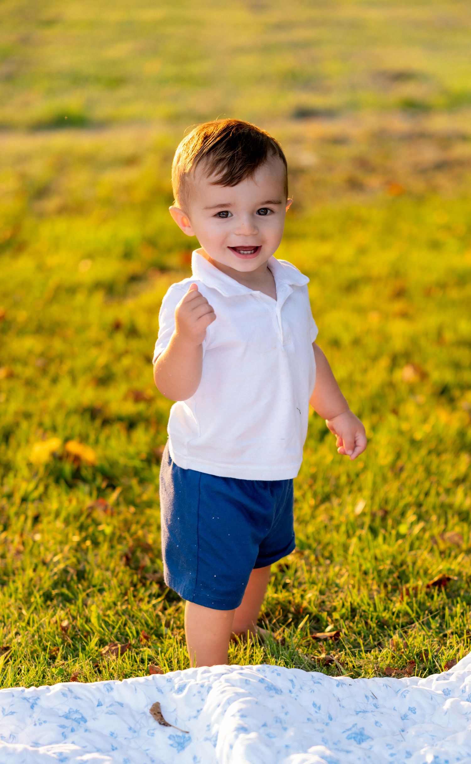 Smiling toddler boy in white polo shirt and navy shorts standing on grass in sunny outdoor setting.