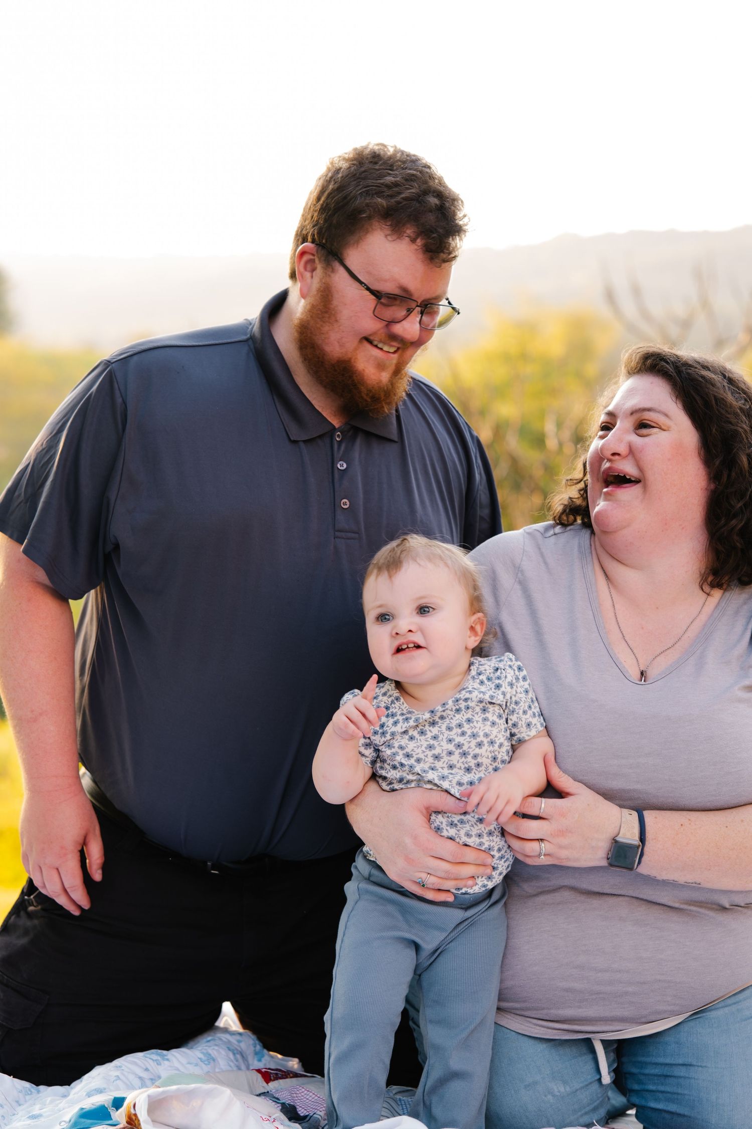 A happy family with a bearded dad, laughing mom, and cute baby enjoy an outdoor picnic at golden hour.